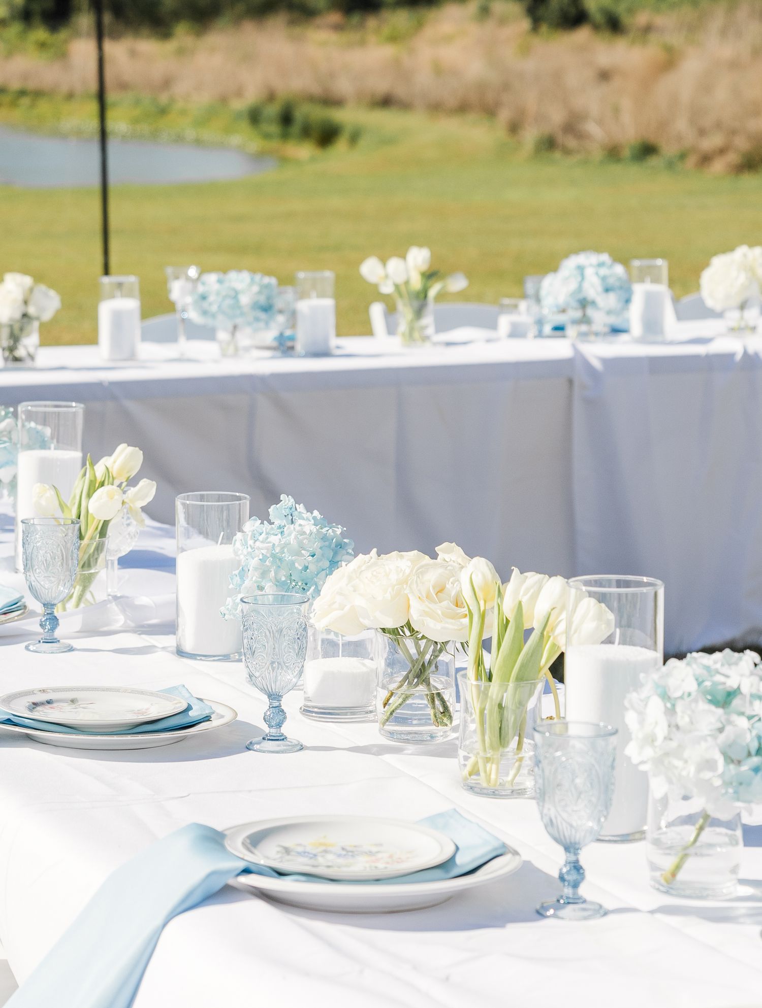Elegant outdoor wedding reception tables with white tulips, blue hydrangeas, blue goblets, candles, and white linens by a pond.