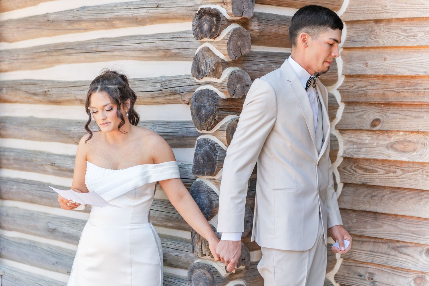 Bride and groom hold hands around a log cabin wall during an emotional first look reading letters.