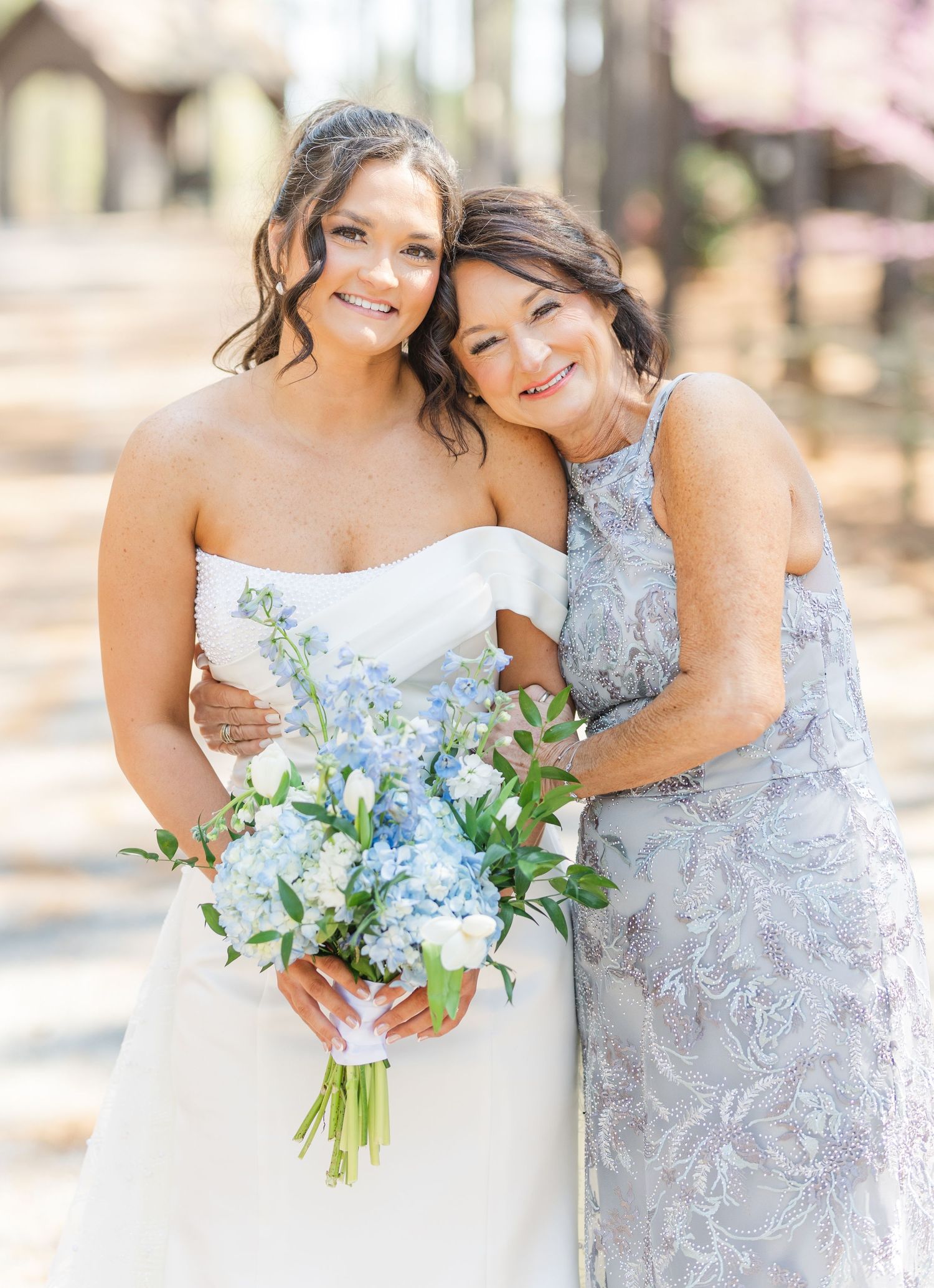 Bride in strapless white gown holding blue hydrangea bouquet with mother in silver dress.