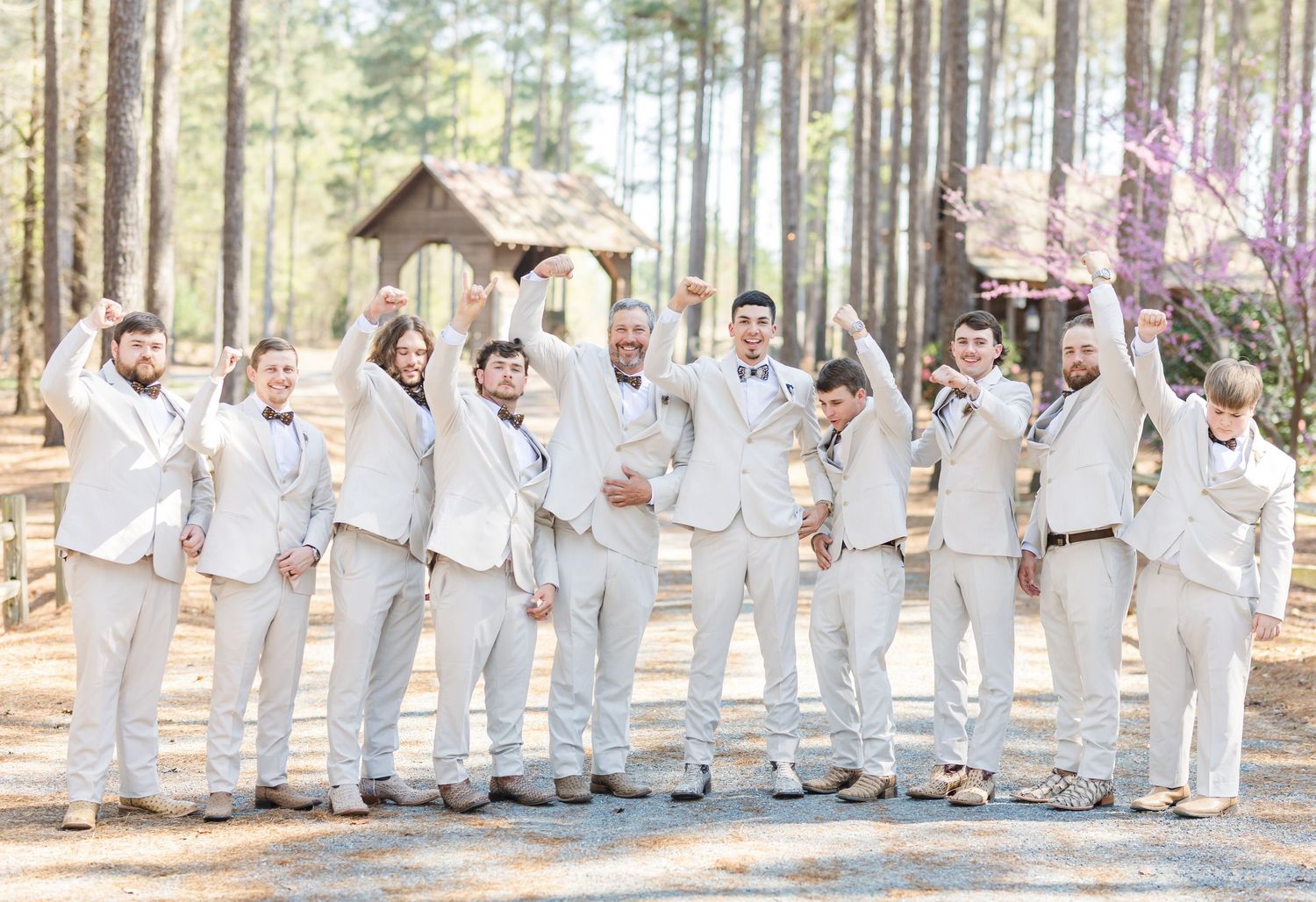 Large groomsmen party in matching cream suits cheering with arms raised at outdoor pine tree wedding.