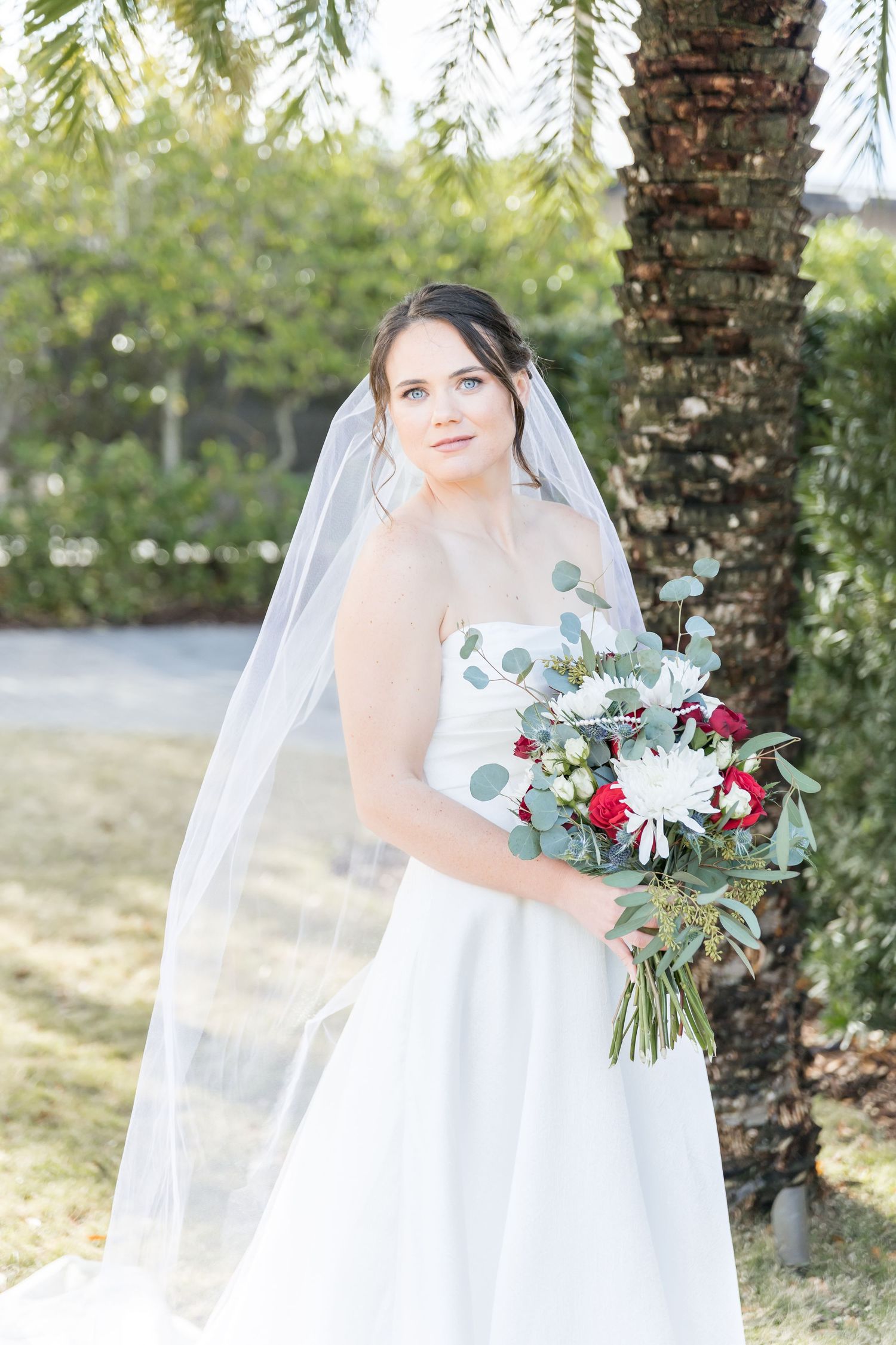 A bride in a white wedding dress poses outdoors holding a bouquet with red roses and white flowers.