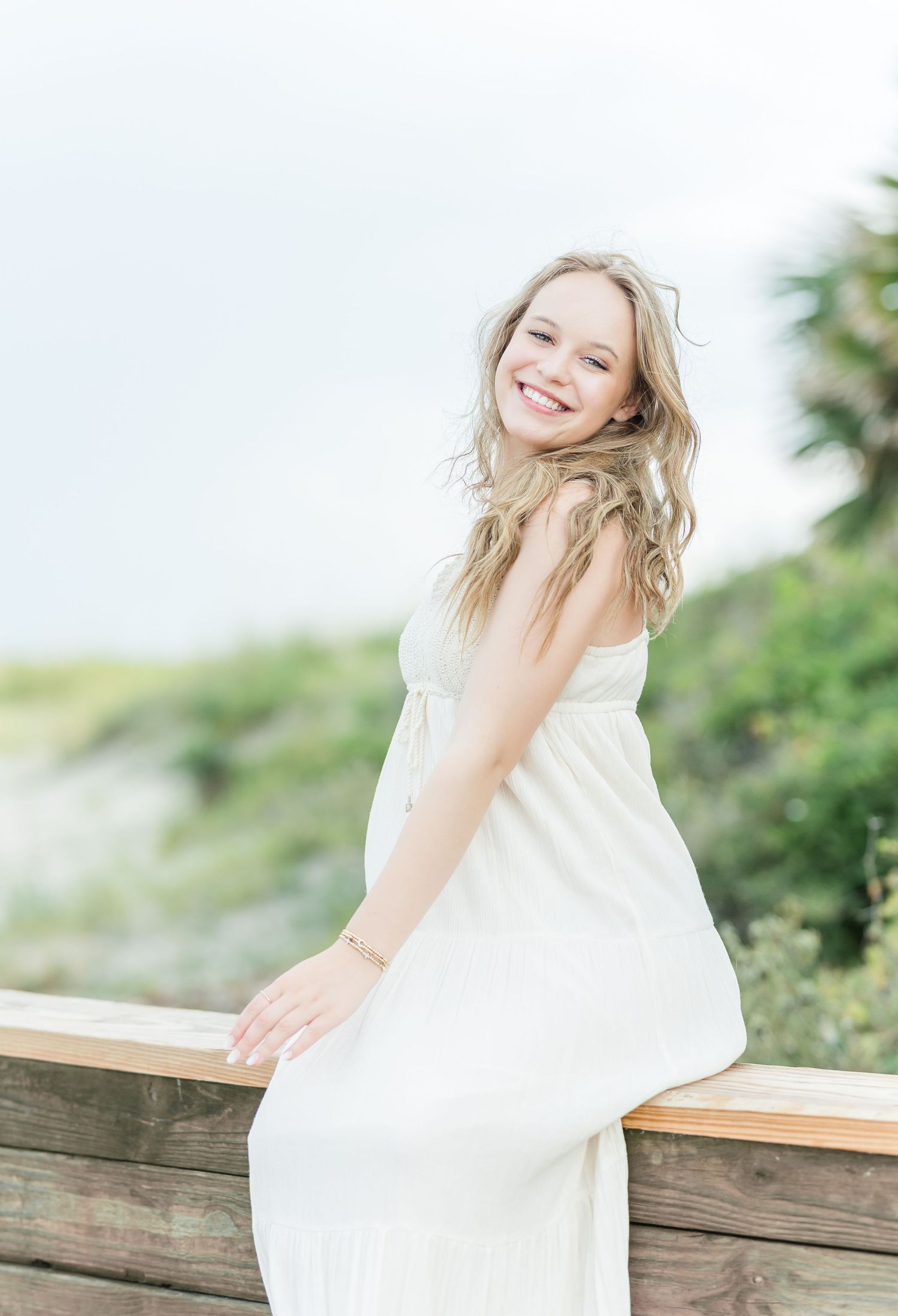 White sundress flows in the breeze as someone poses against a wooden railing by the coast.