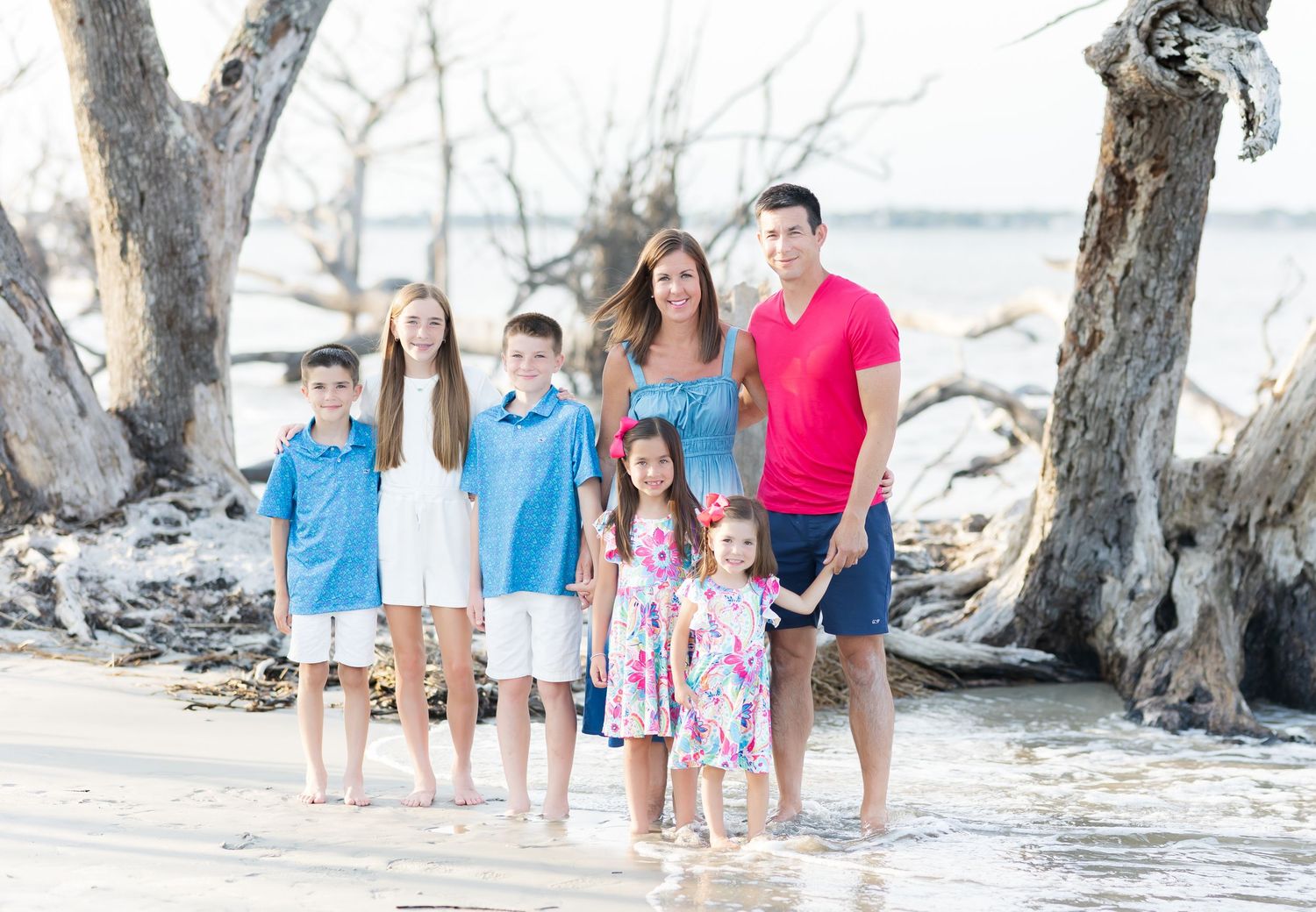 A family stands together on a sunny beach with driftwood and ocean views in the background.