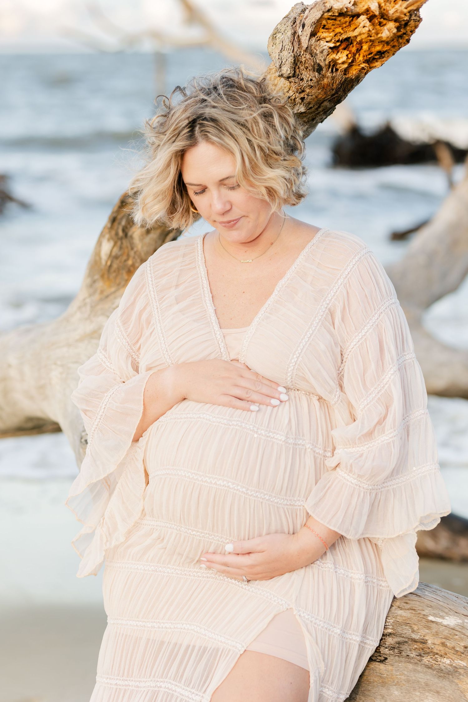 Serene maternity portrait showing woman in cream dress resting against weathered driftwood on beach.