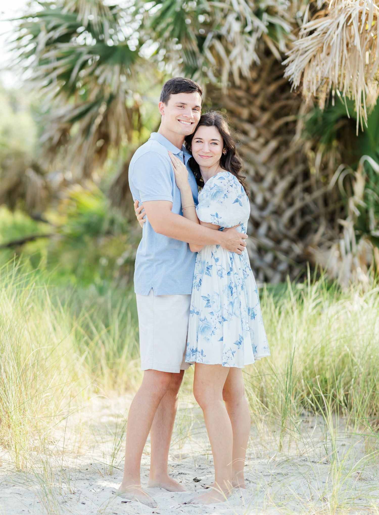 Romantic couple in coordinating blue and white outfits embrace on a sandy beach with palm trees in background.