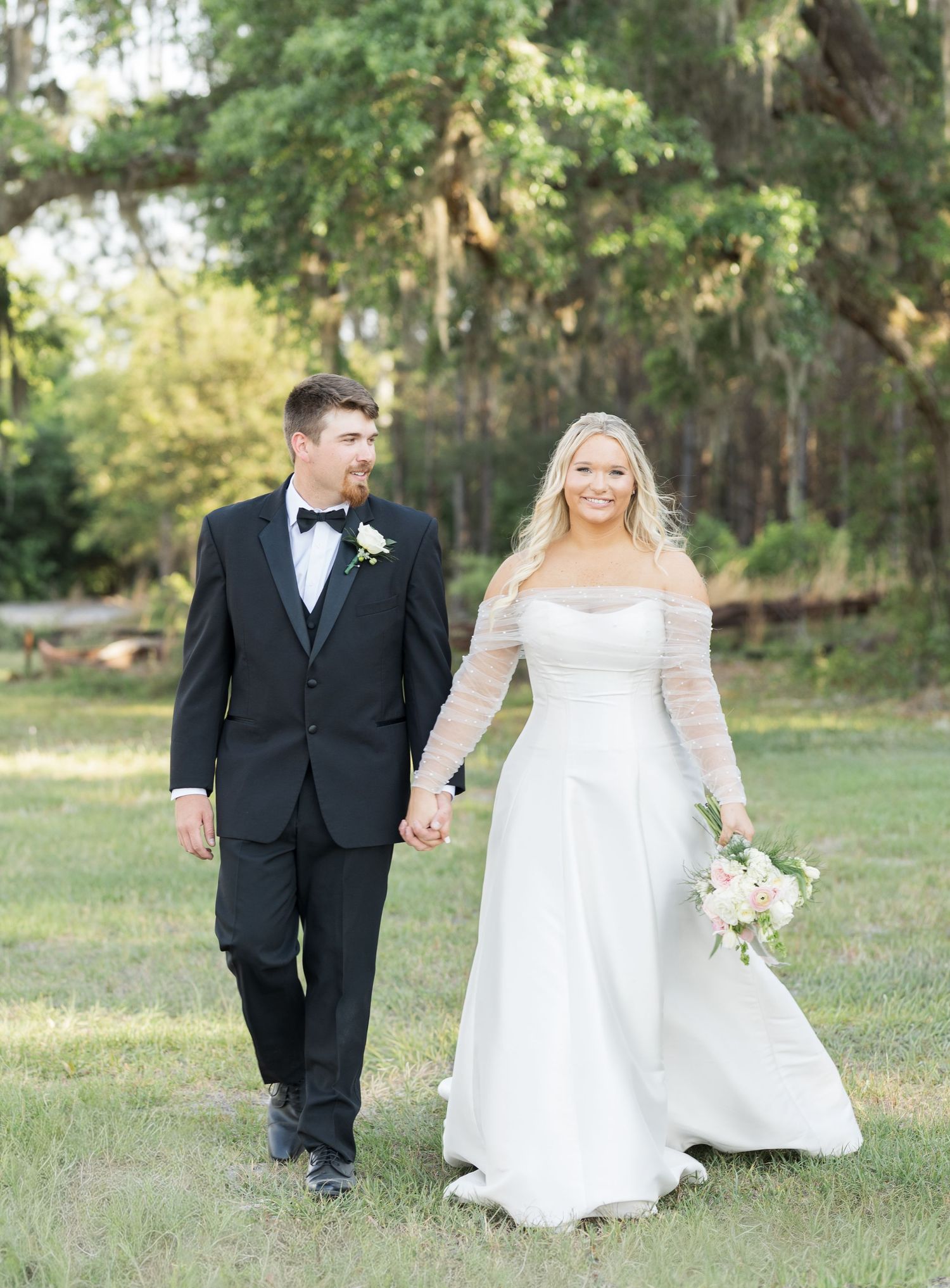 Bride and groom walk hand-in-hand outdoors, she in a white off-shoulder gown, he in a classic black tuxedo.