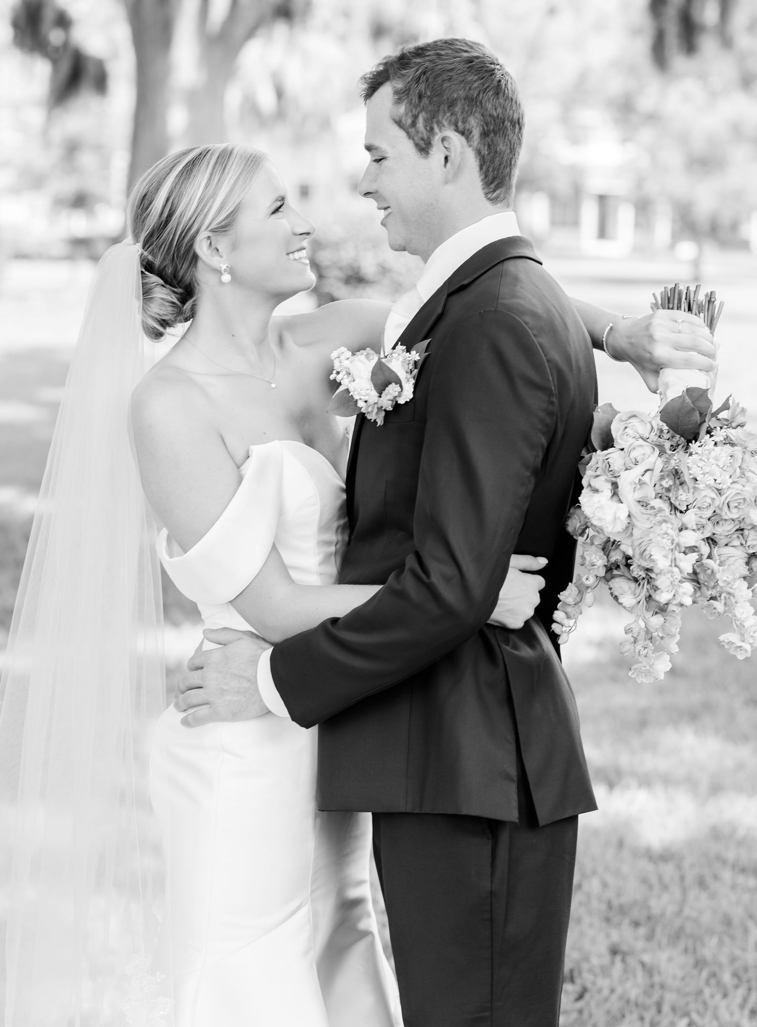 Black and white photo of a smiling bride and groom embracing outdoors, bride holding bouquet and wearing veil.
