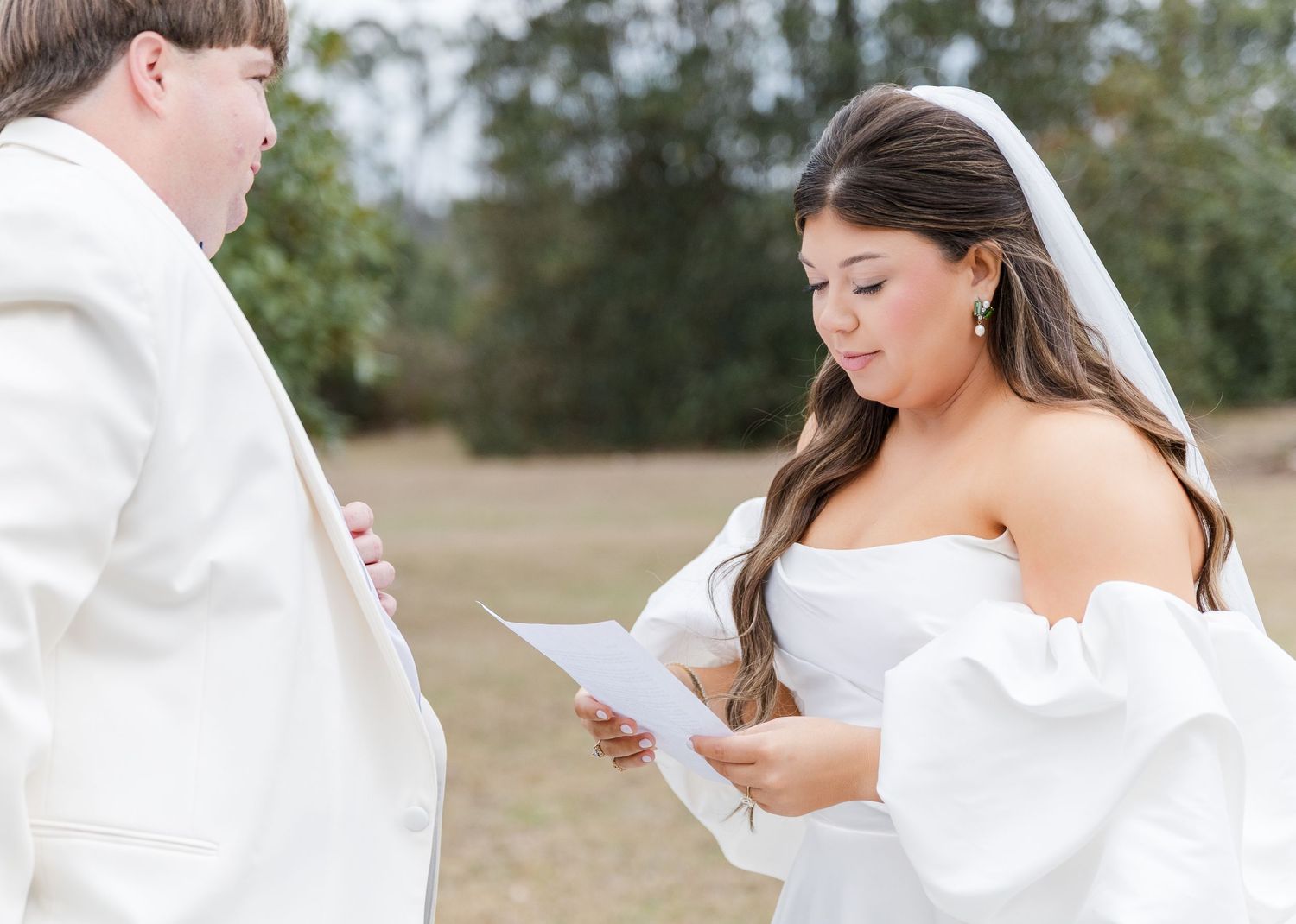 Bride in white off-shoulder gown reads vows from paper during outdoor wedding ceremony.