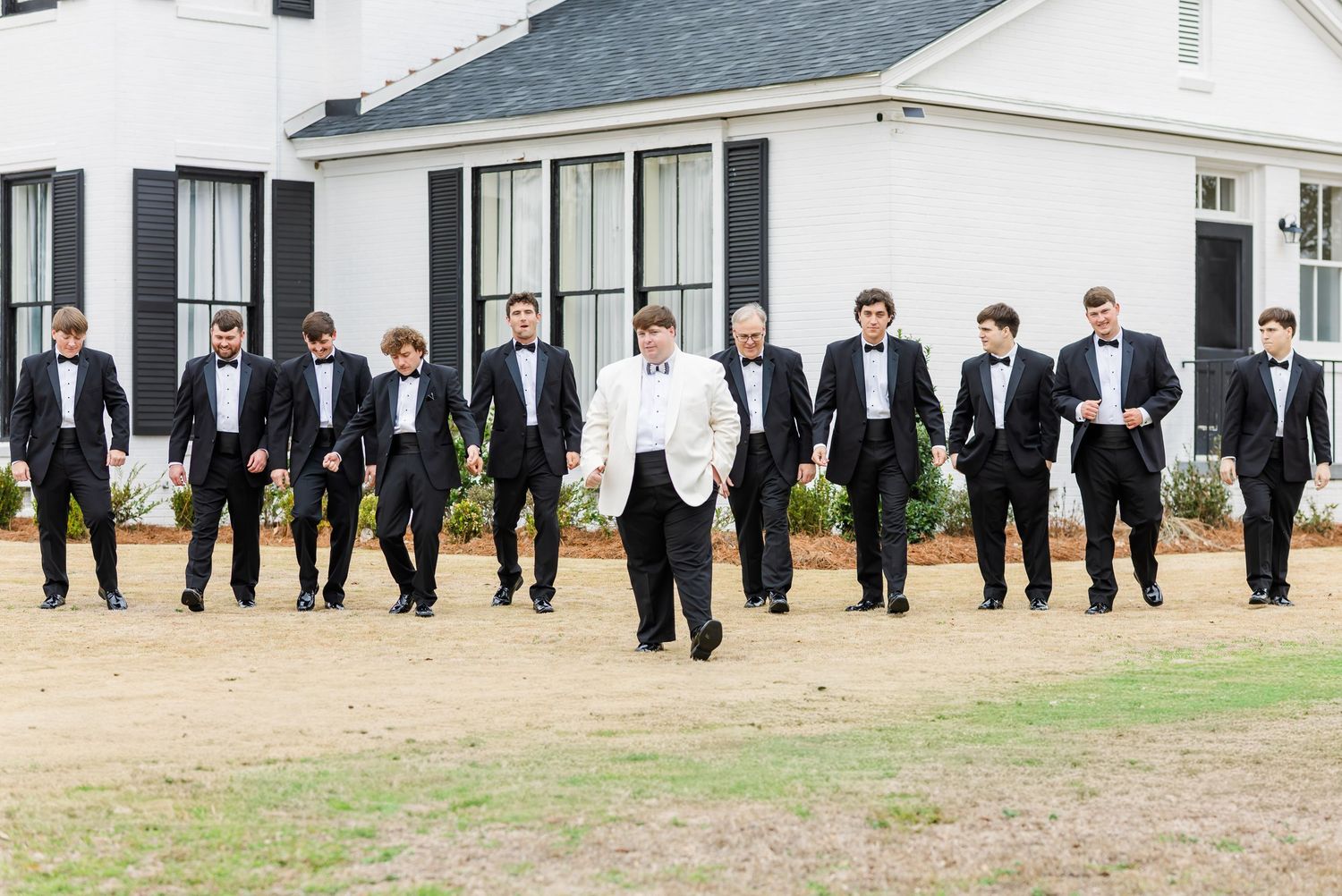 Groom in white tuxedo leads groomsmen in black tuxedos walking across lawn at wedding venue.