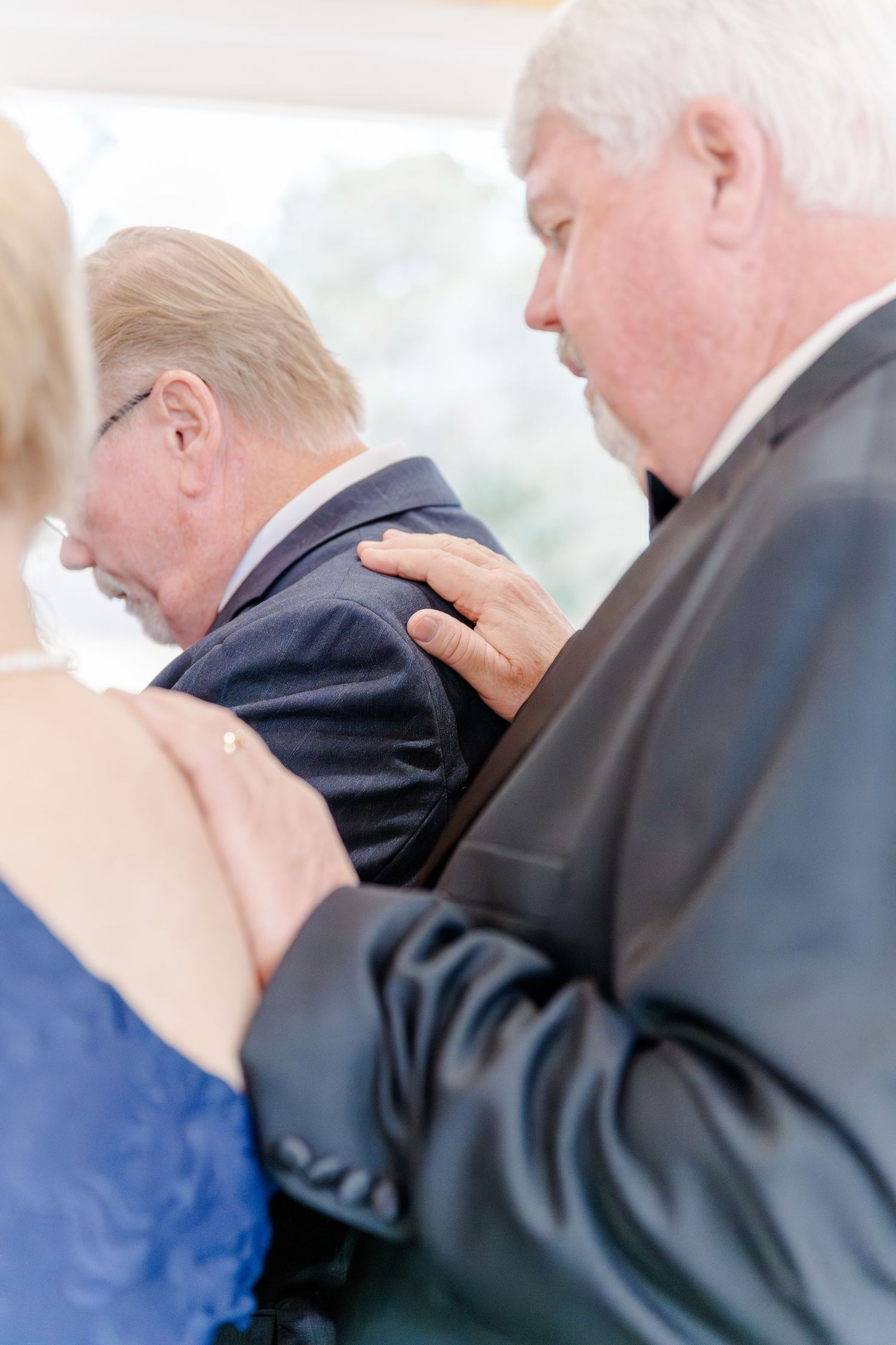 Two older men in suits bowing heads in prayer at a wedding ceremony, with a woman's hand on one's shoulder.