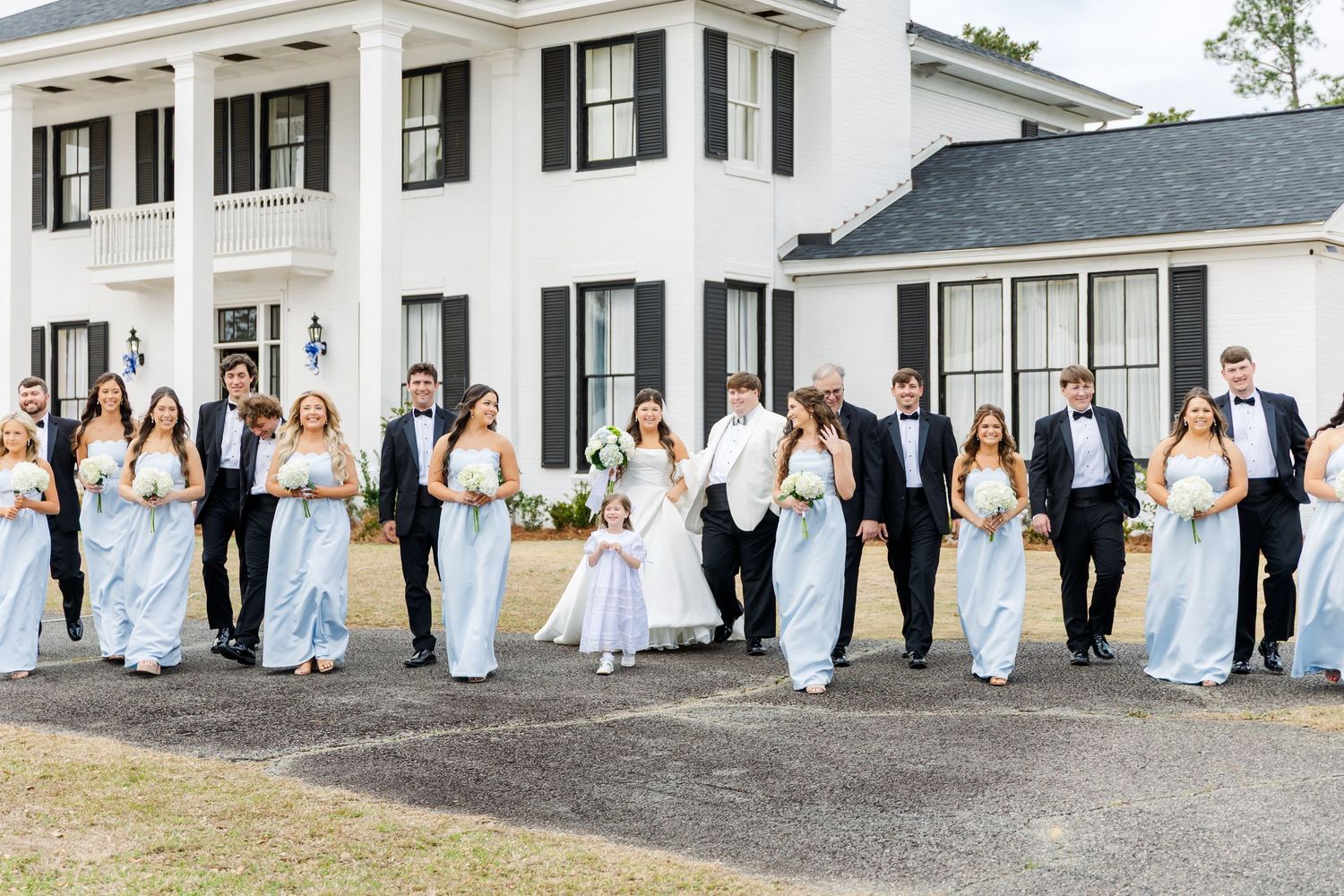 Full wedding party in light blue and black formal attire walking outside white wedding venue.