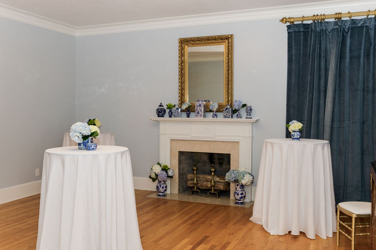 Elegant wedding reception room with white cocktail tables, fireplace, gold mirror, and blue floral arrangements.