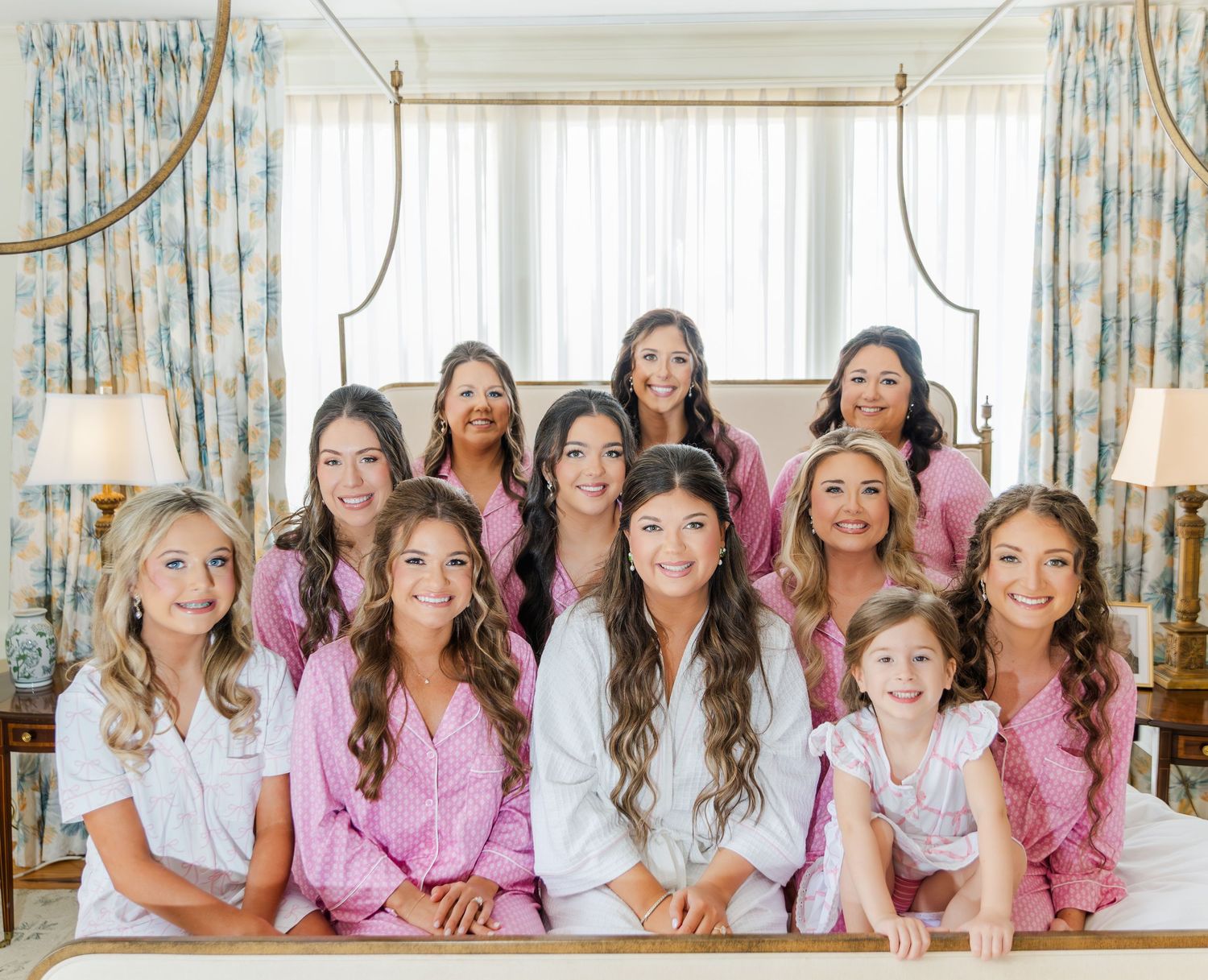 Bride and bridesmaids in pink satin robes posing together on bed during wedding morning preparations.