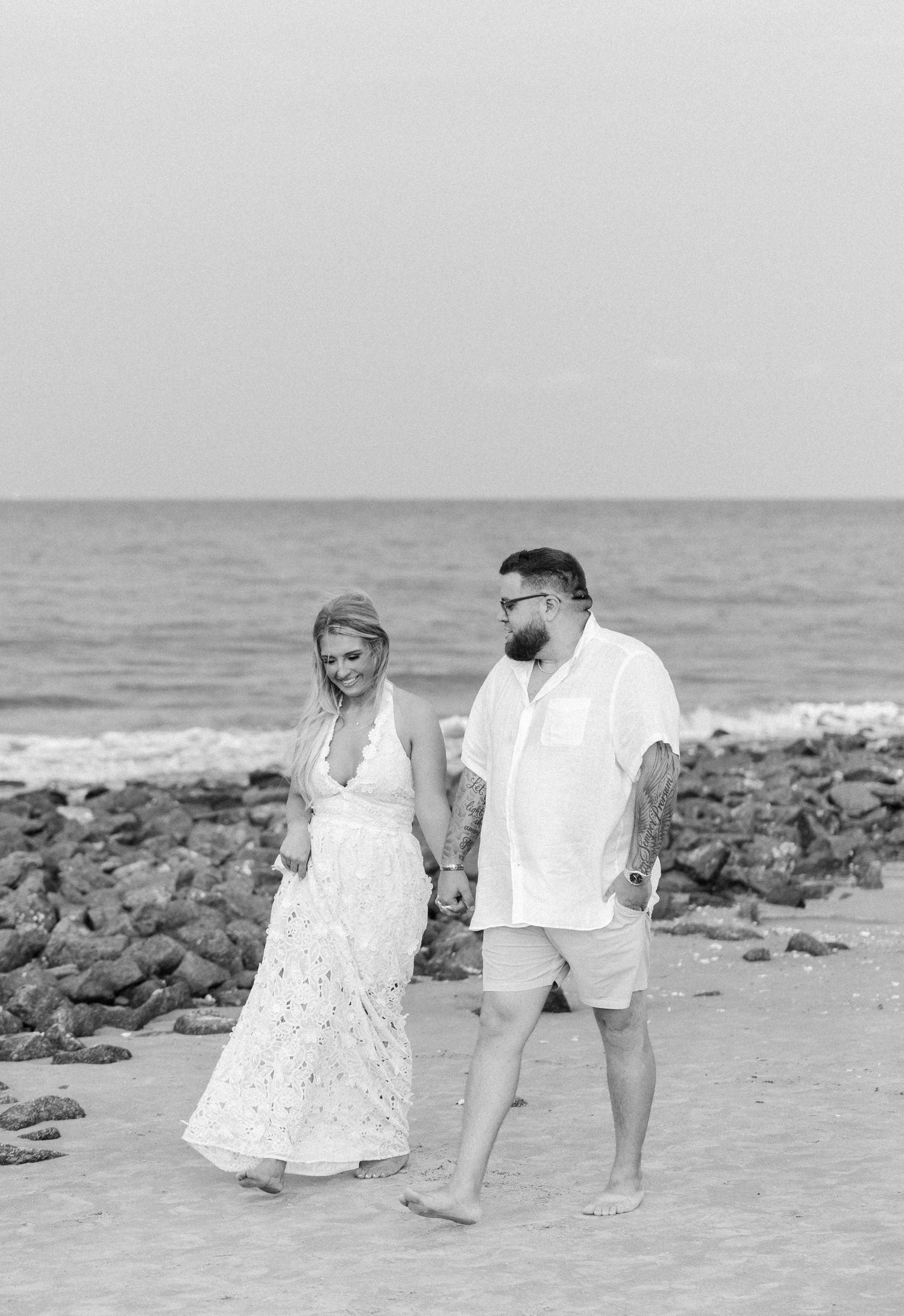A couple walks hand-in-hand on a rocky beach, woman in white dress, man in linen shirt, black and white photo.