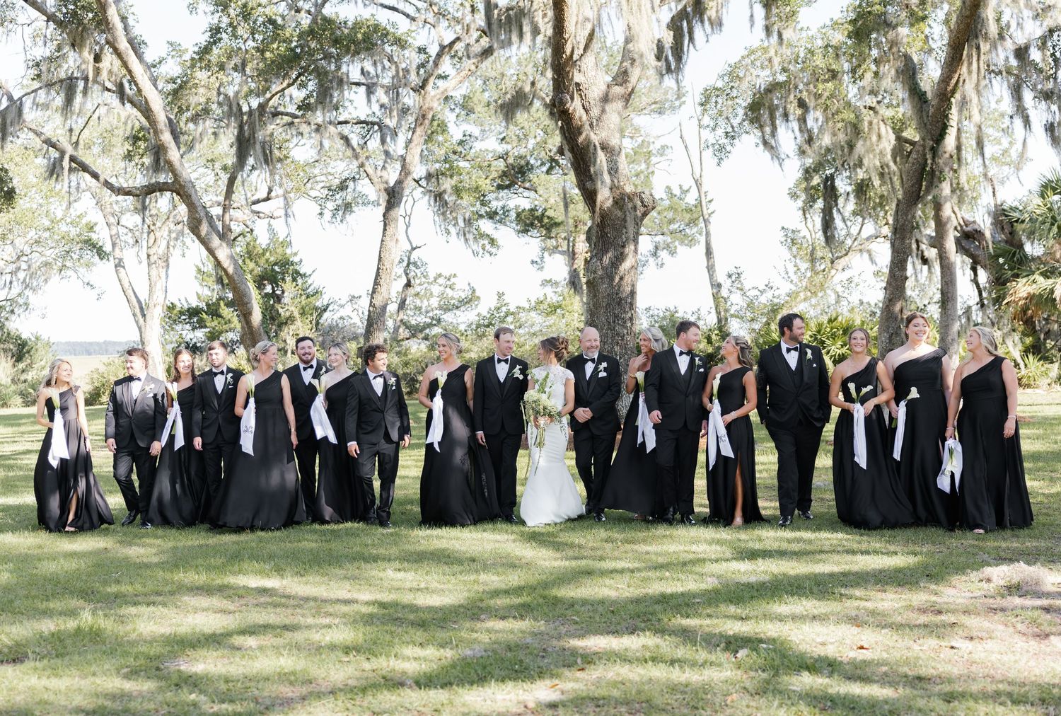 Wedding party in black attire poses outdoors under Spanish moss-draped oak trees on a sunny day.