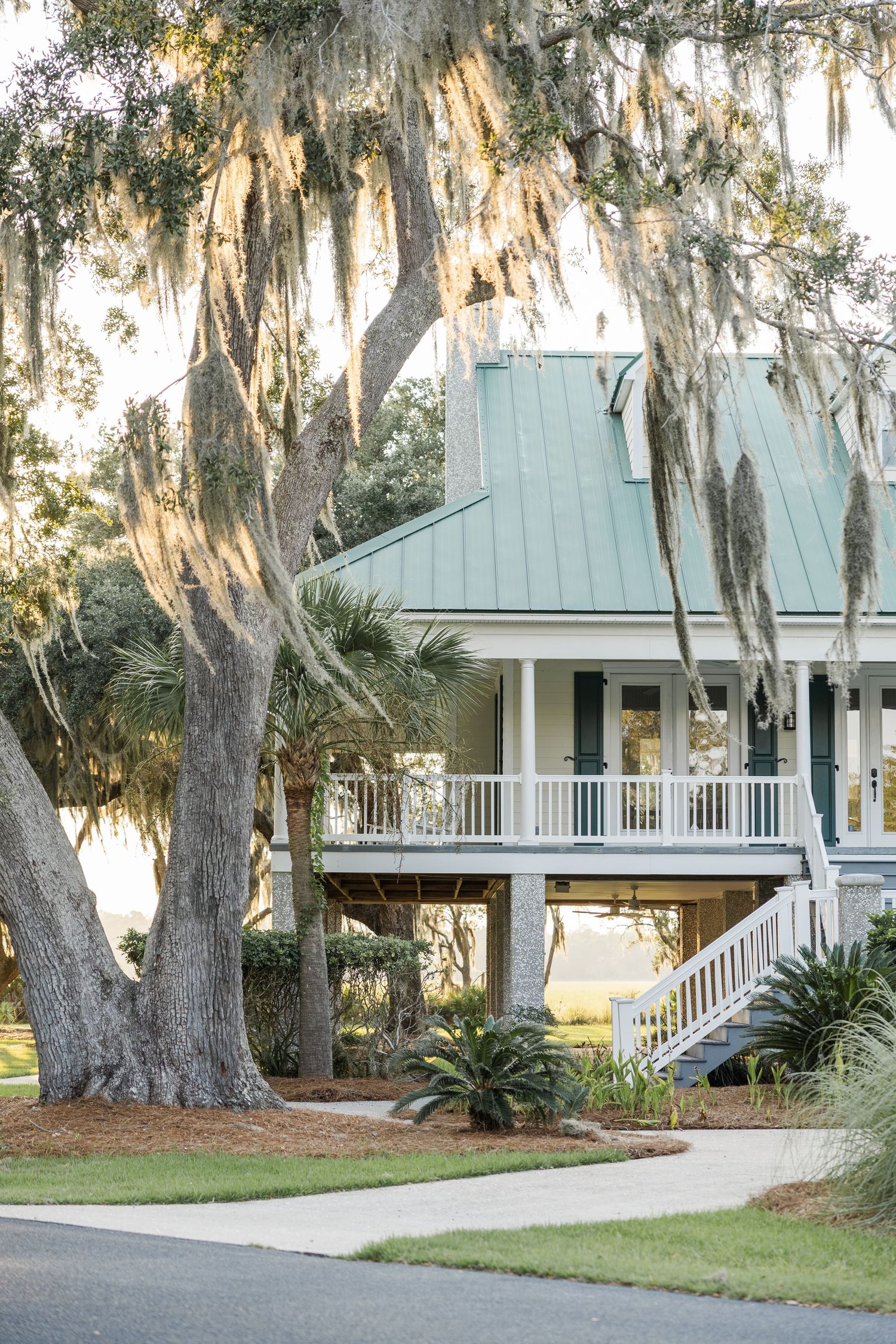 Southern coastal home with teal metal roof, white railings, and Spanish moss-draped oak trees in warm evening light.