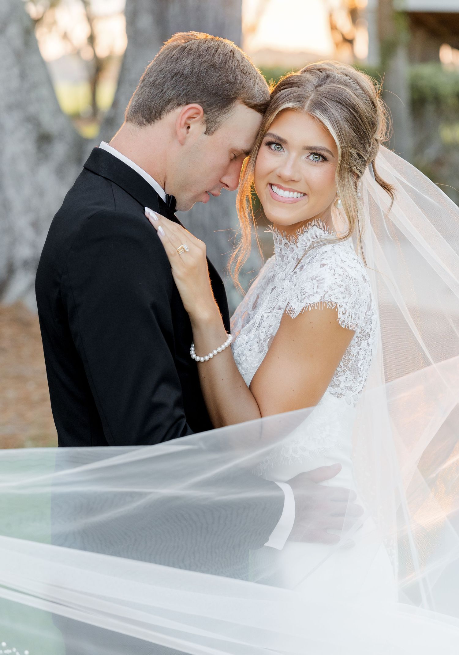 Bride in lace gown and veil smiles over shoulder while groom in black tuxedo holds her close outdoors.