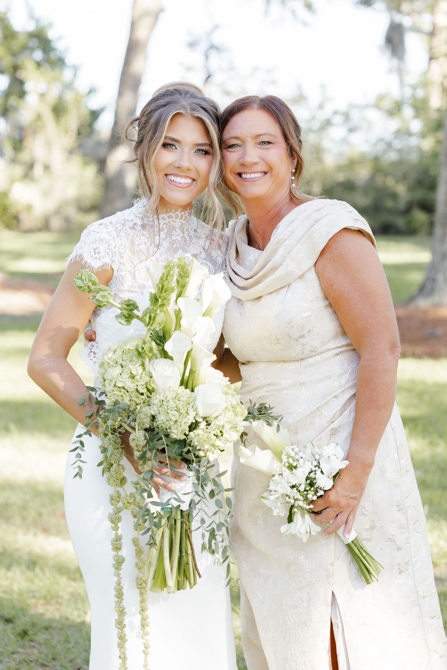 Two women in elegant white gowns holding green and white bouquets pose together outdoors at a wedding celebration.