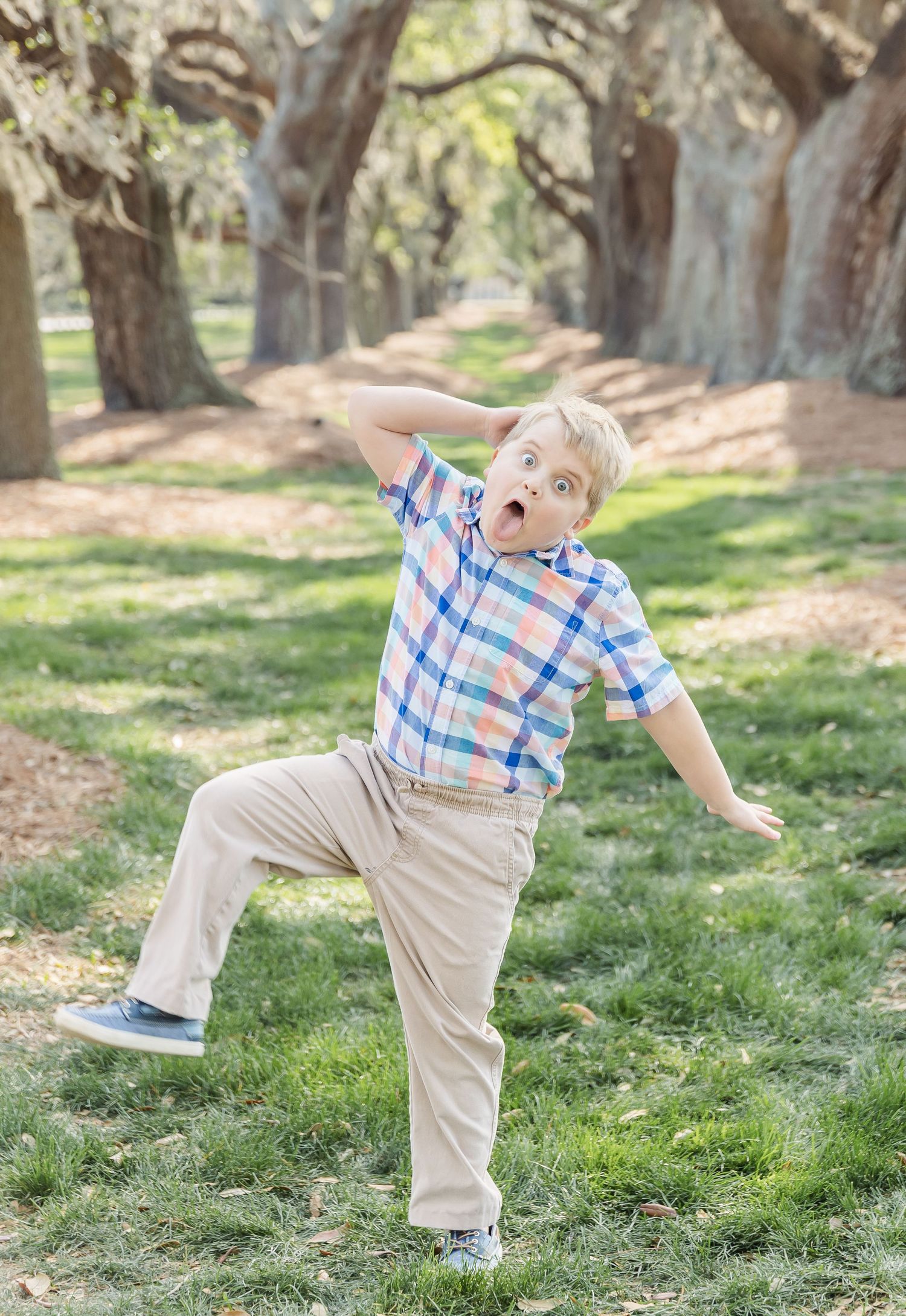 Playful child in plaid shirt striking silly pose on tree-lined path in park.