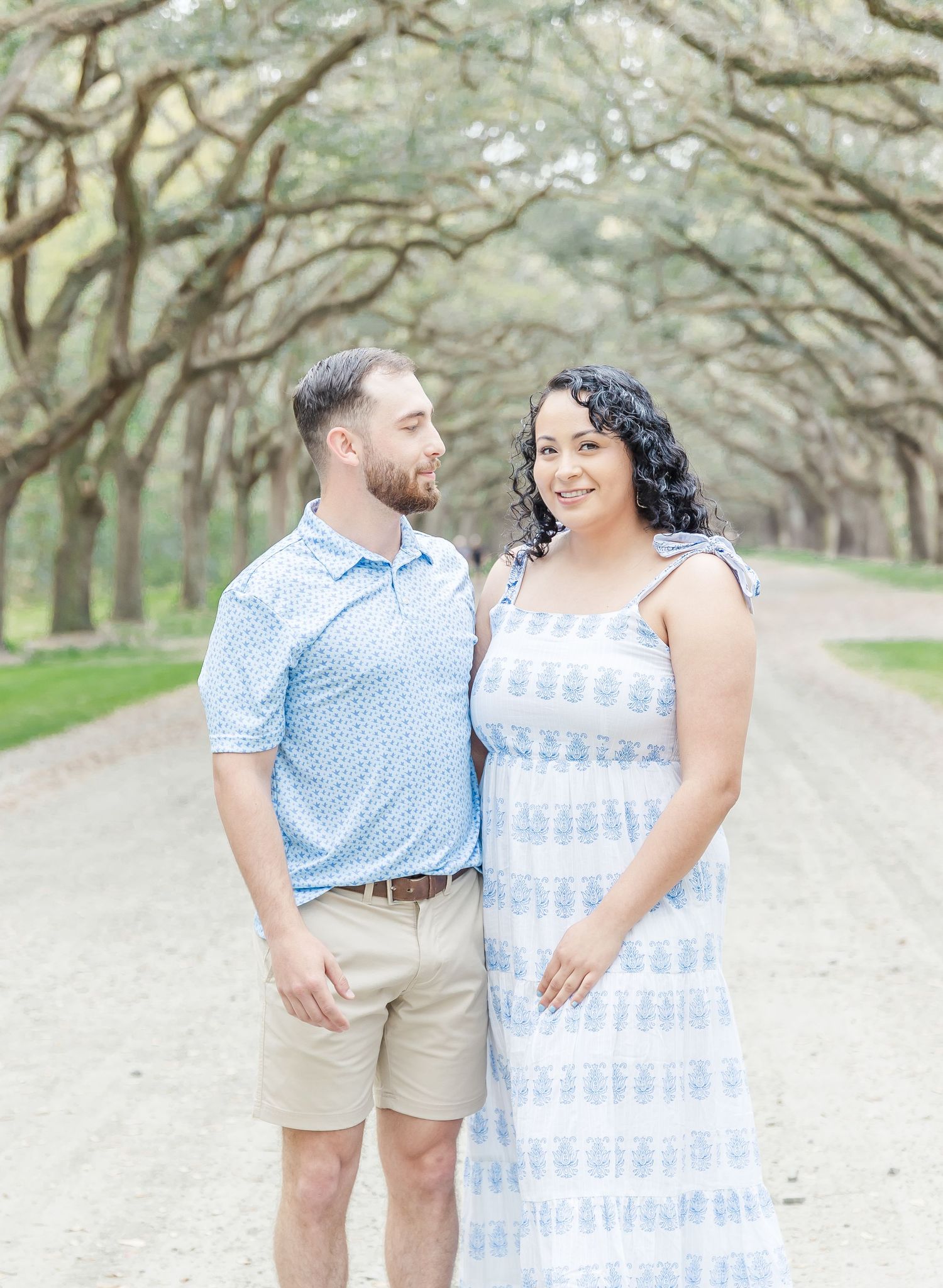 Couple in blue and white attire share tender moment under arching oak trees.