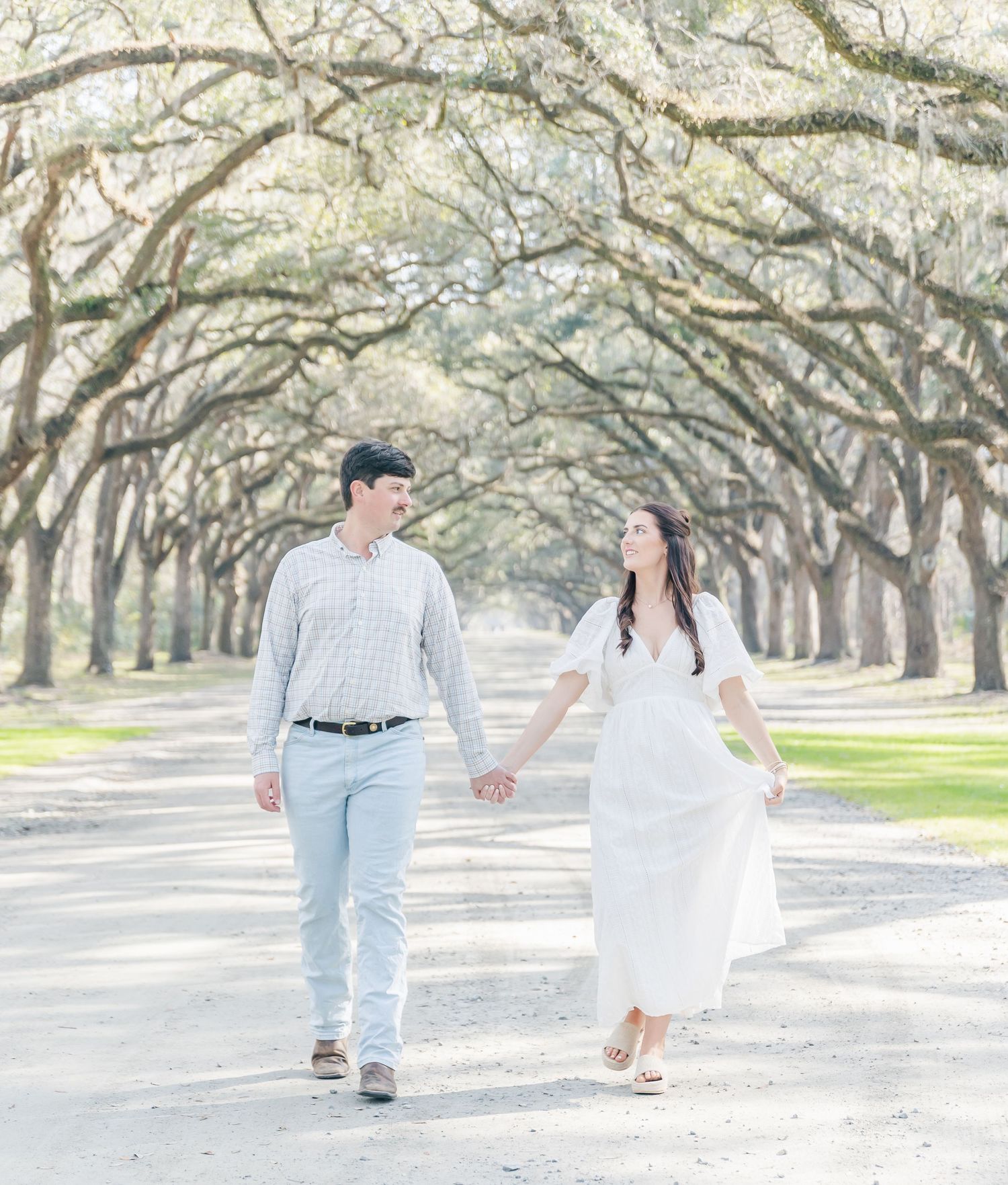 A couple walk hand in hand down a dreamy oak-lined path in light, airy clothing.
