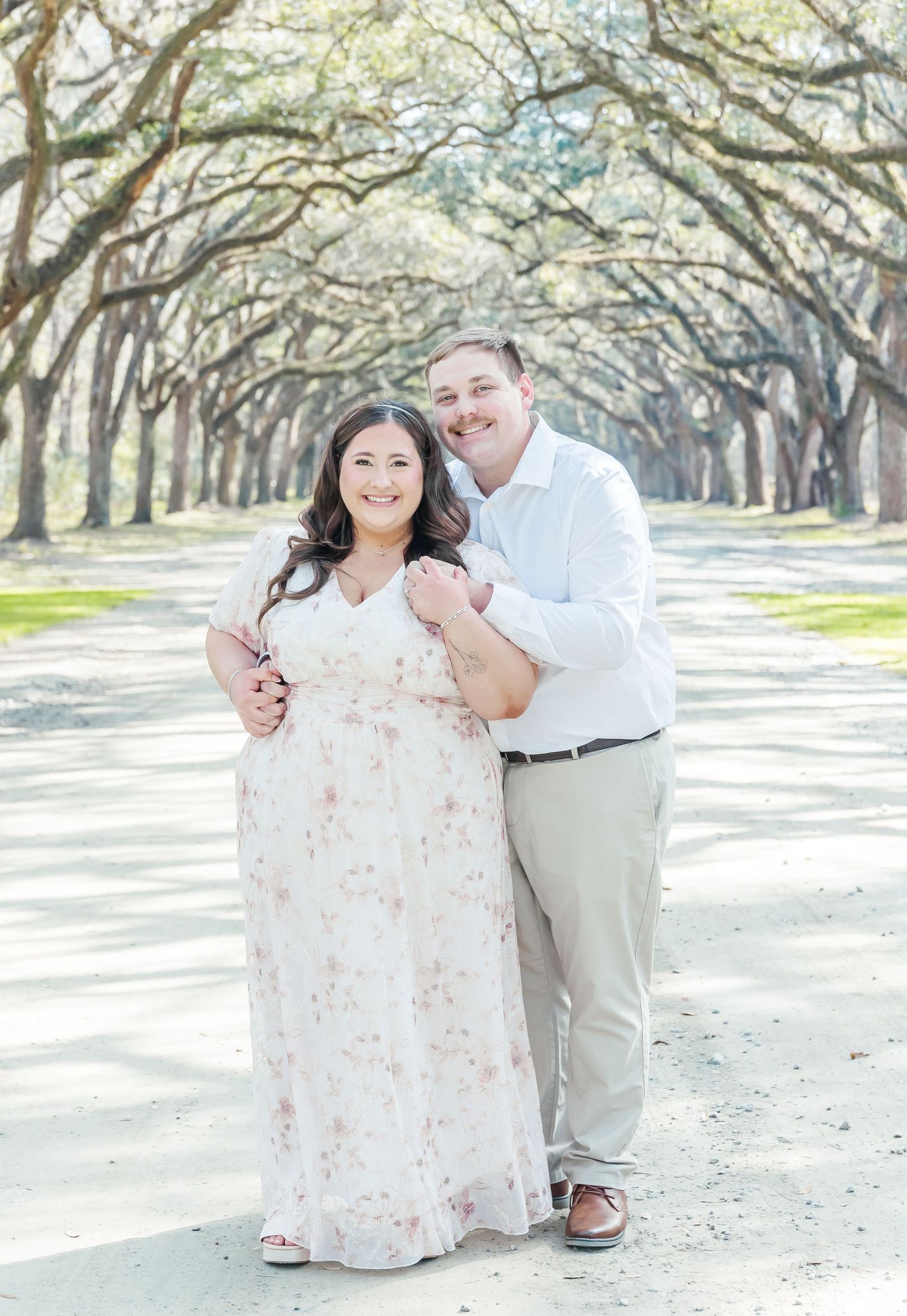 Couple smiling for engagement photos under a natural archway of oak trees.