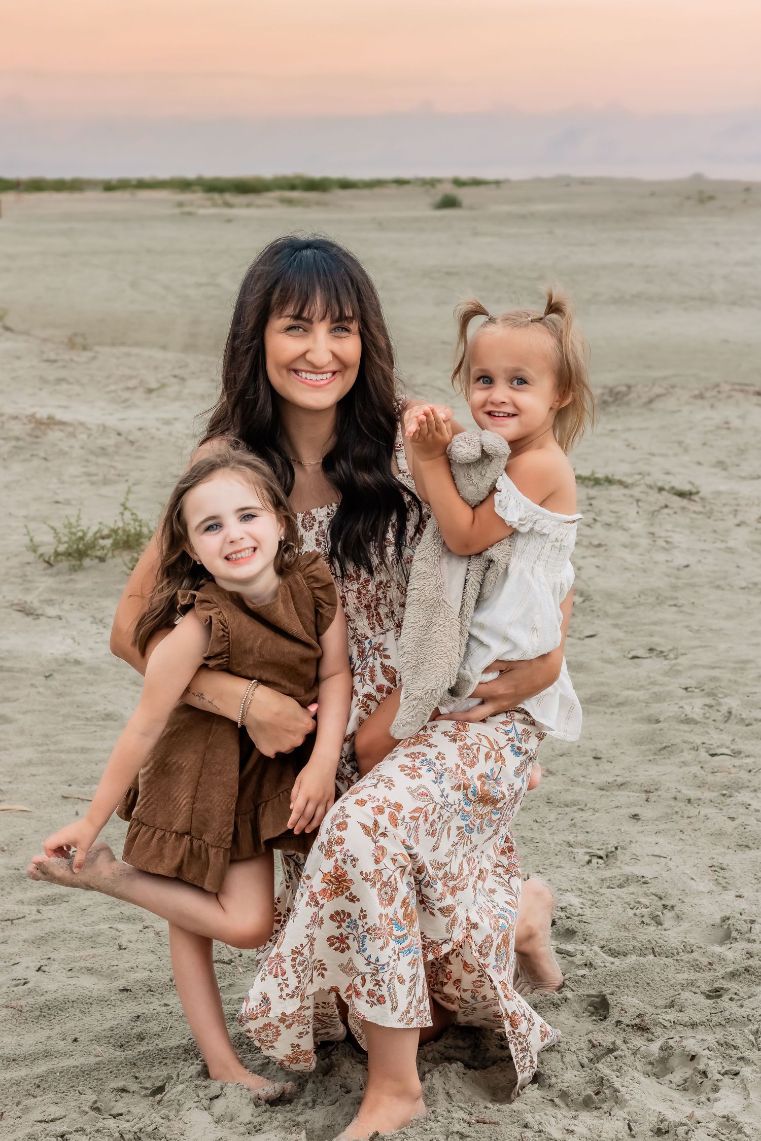Mother and two daughters pose on sandy beach at sunset in flowing dresses.