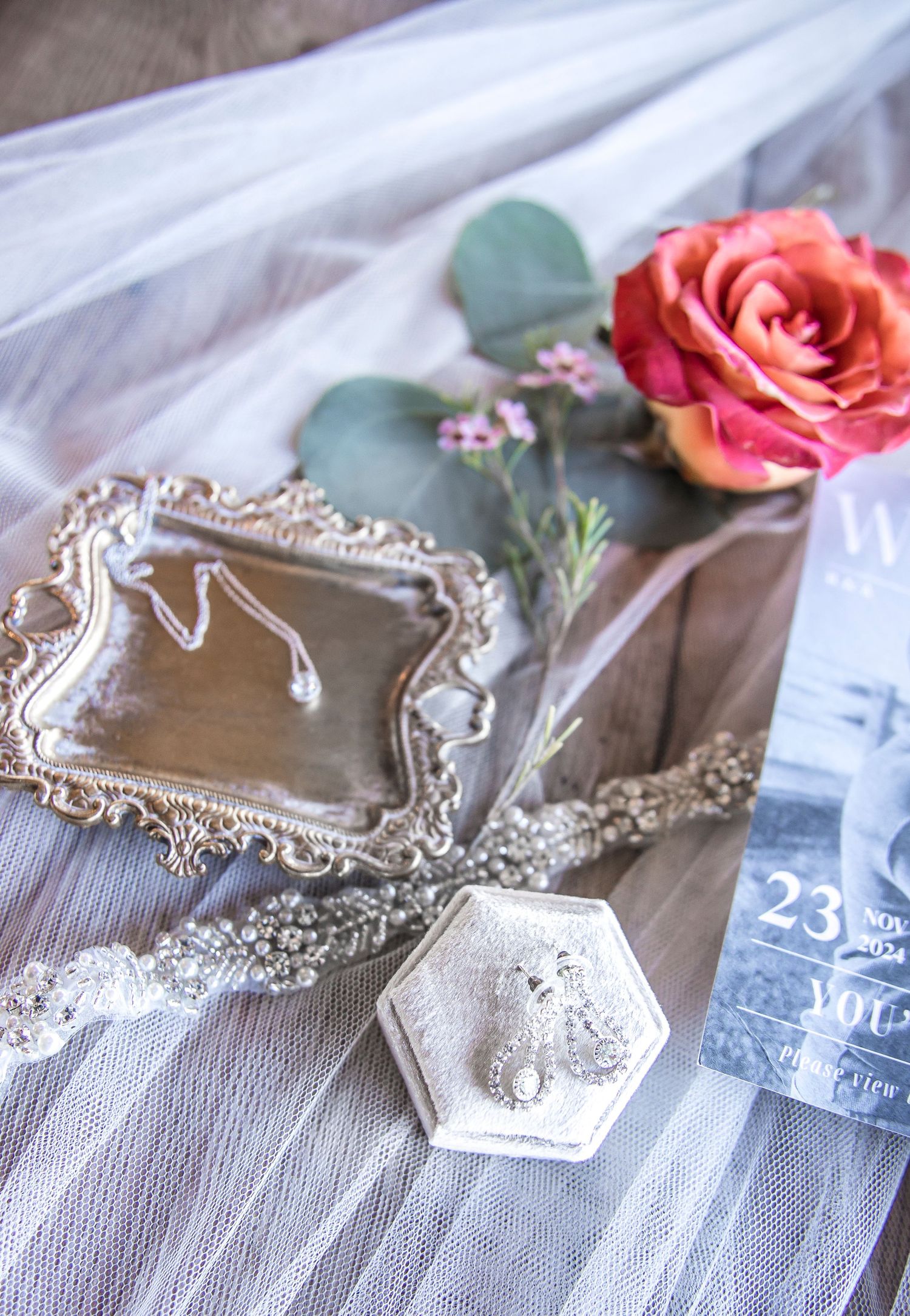 Romantic wedding details with coral rose, vintage mirror, and white ring box on wood surface.