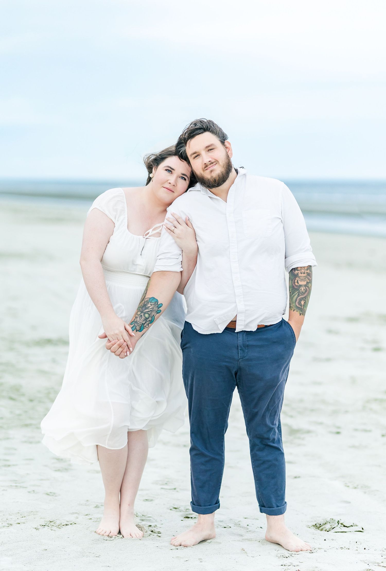 Couple poses together barefoot on a serene beach in casual wedding attire.