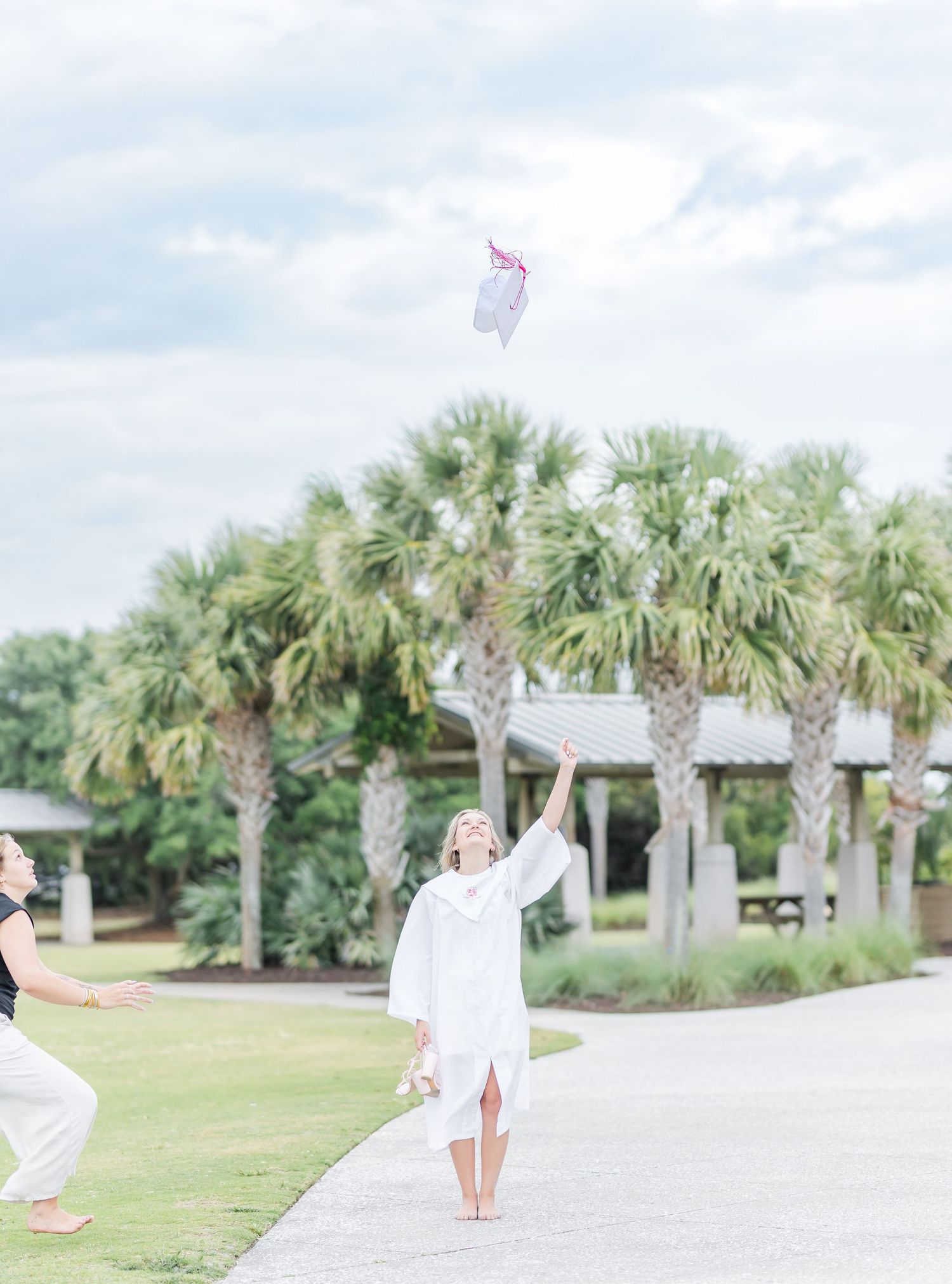 Graduate celebrates by tossing cap in air near tropical landscaping.