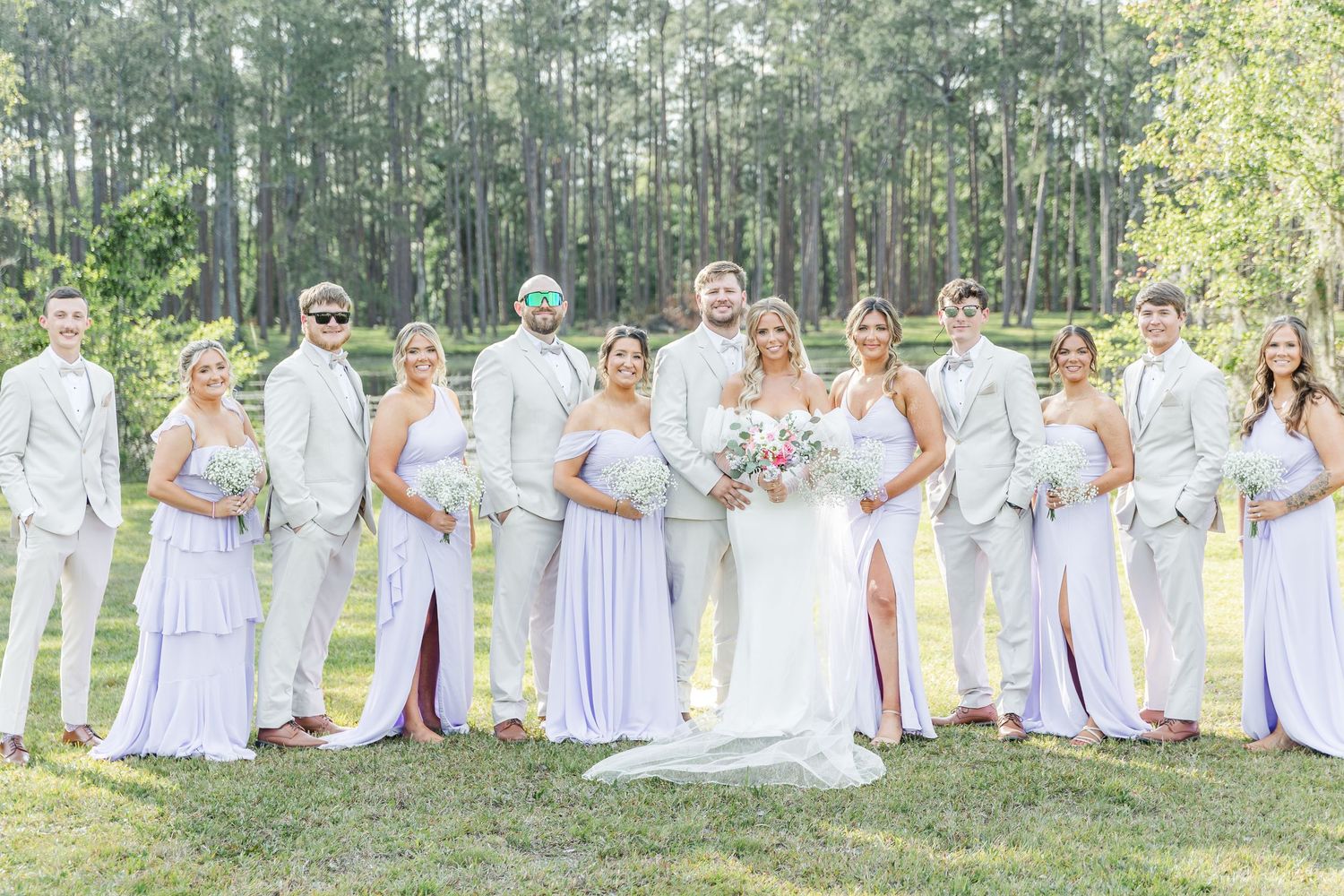 Wedding party in white and lavender attire lined up against a pine forest backdrop.