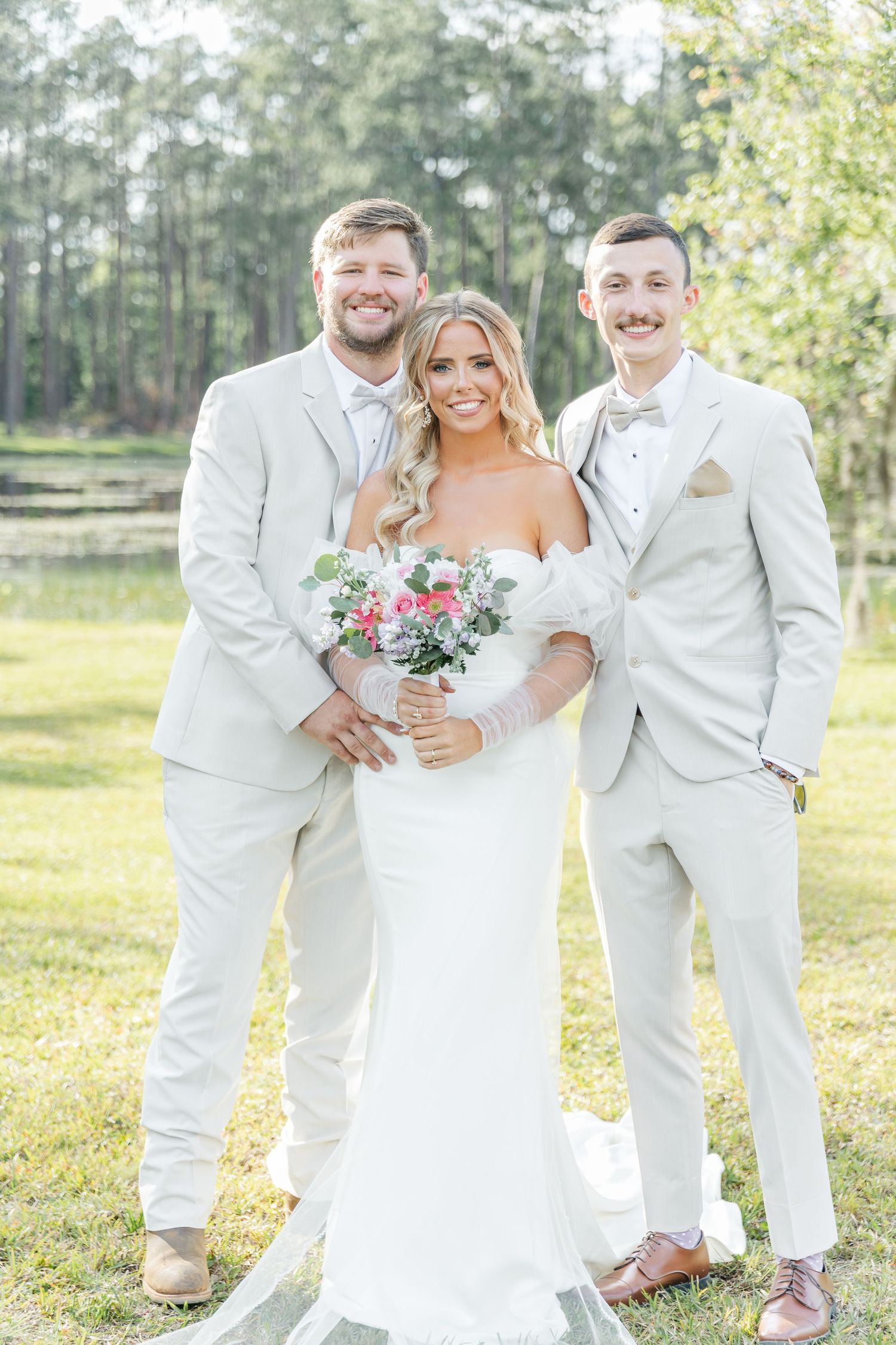 Three people in crisp white attire smile together on sunny lawn with forest backdrop.