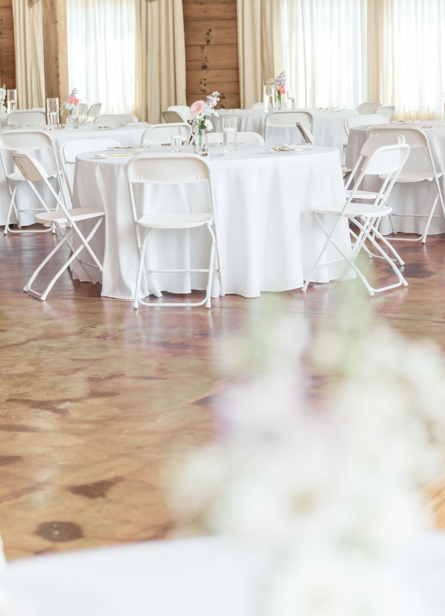Round tables with white linens and folding chairs arranged on polished wooden floors for a wedding reception.