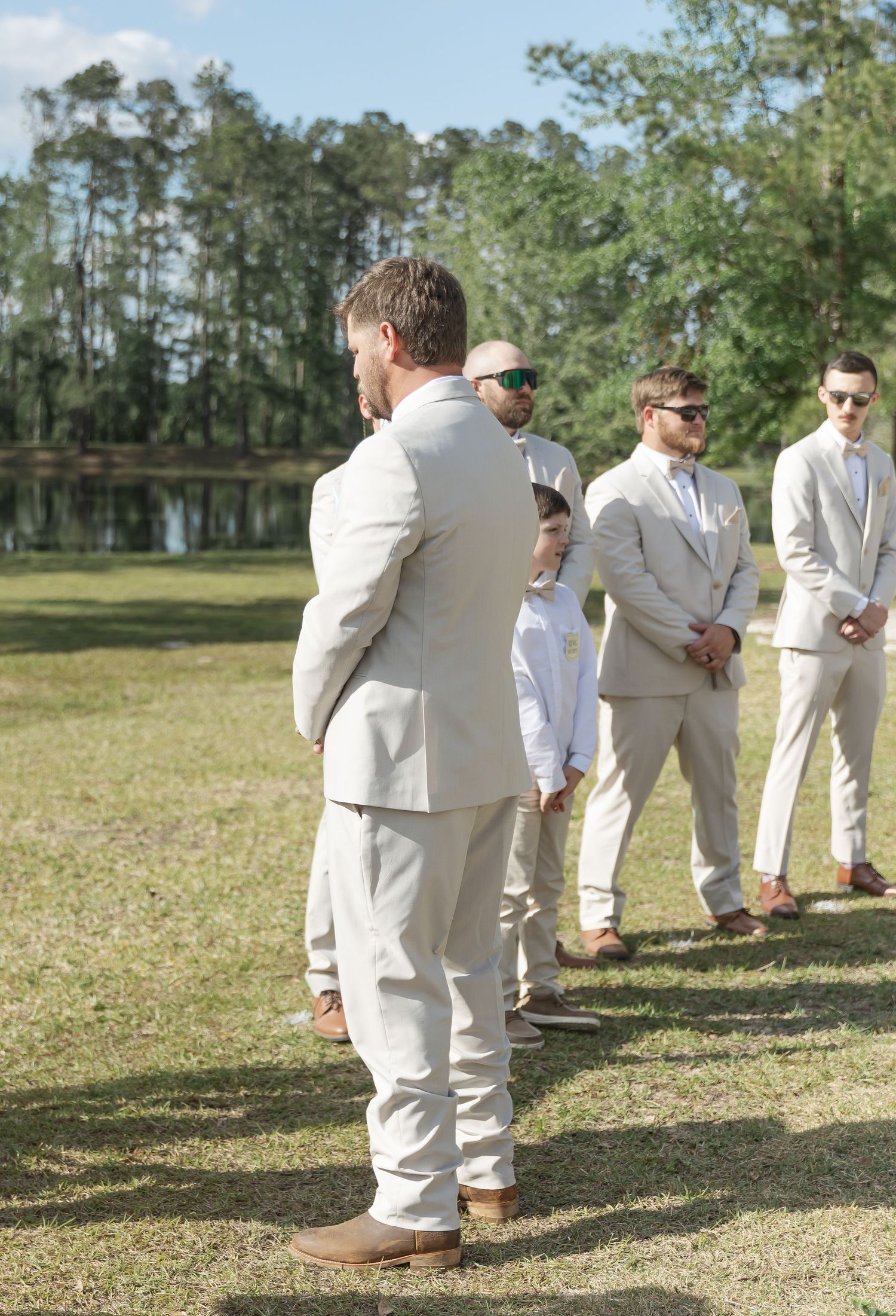 Groomsmen in light colored suits standing on grass near lake during outdoor wedding.