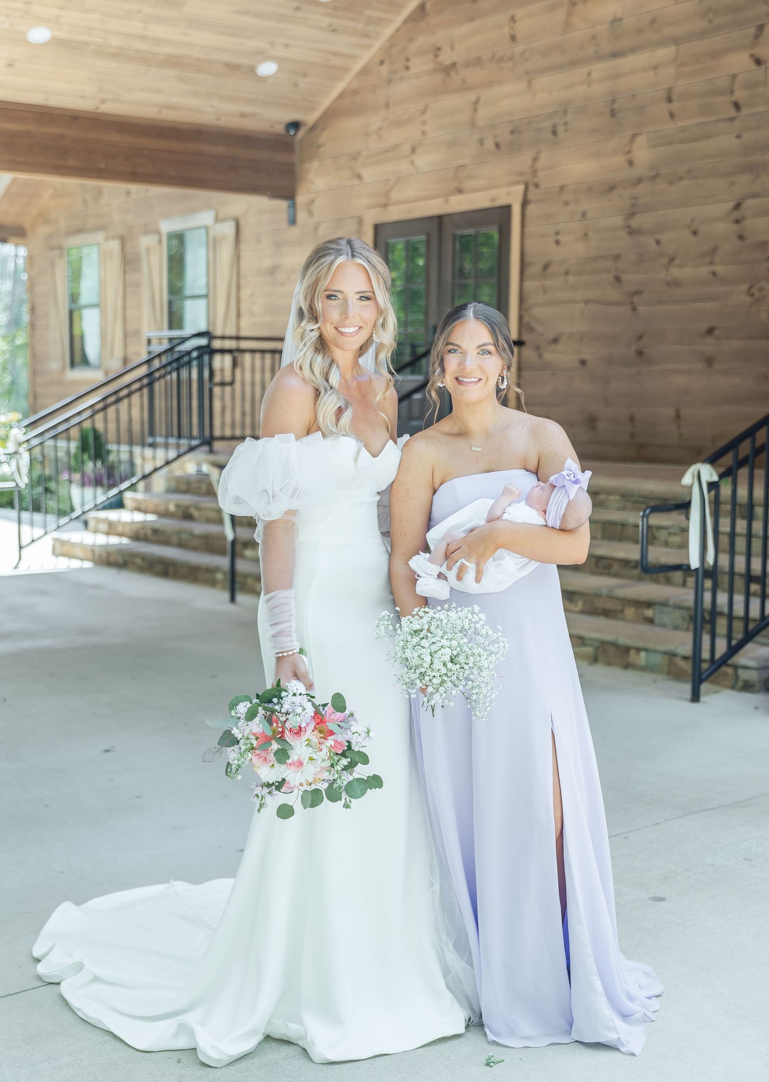 Two women in wedding attire hold infants while posing on wooden porch with bouquets.