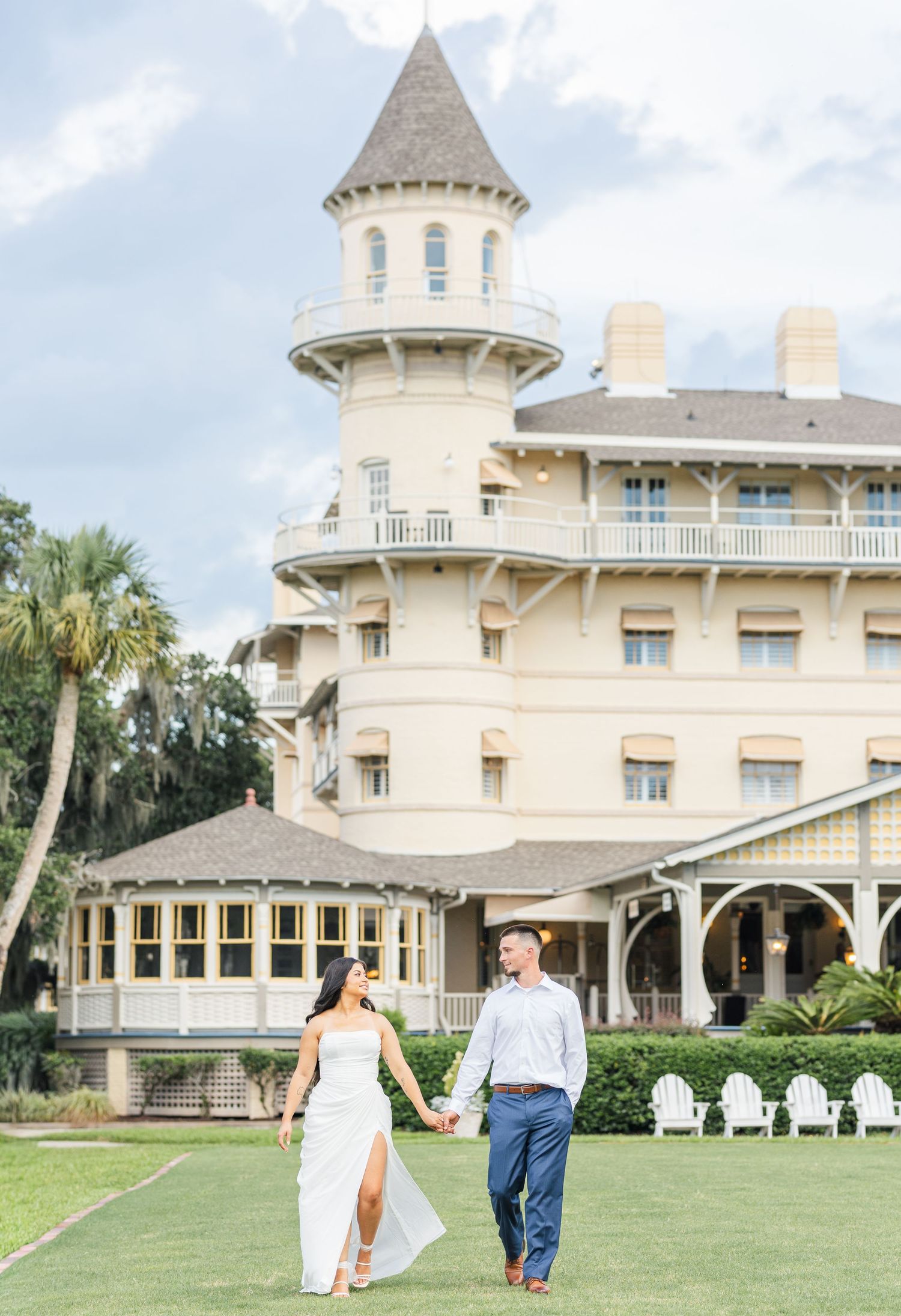 Couple walking hand in hand on lawn before grand yellow Victorian hotel with turret.