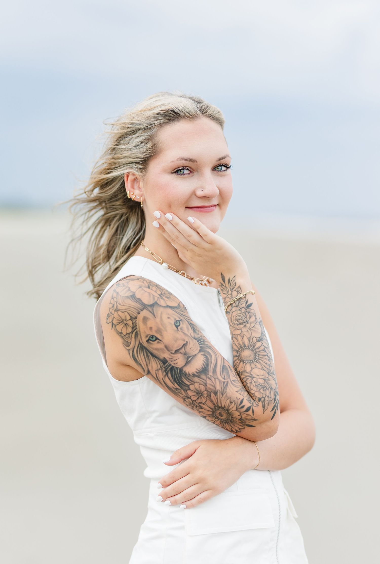 Person in white dress with detailed arm tattoo poses on beach with wind-blown hair in soft natural lighting.