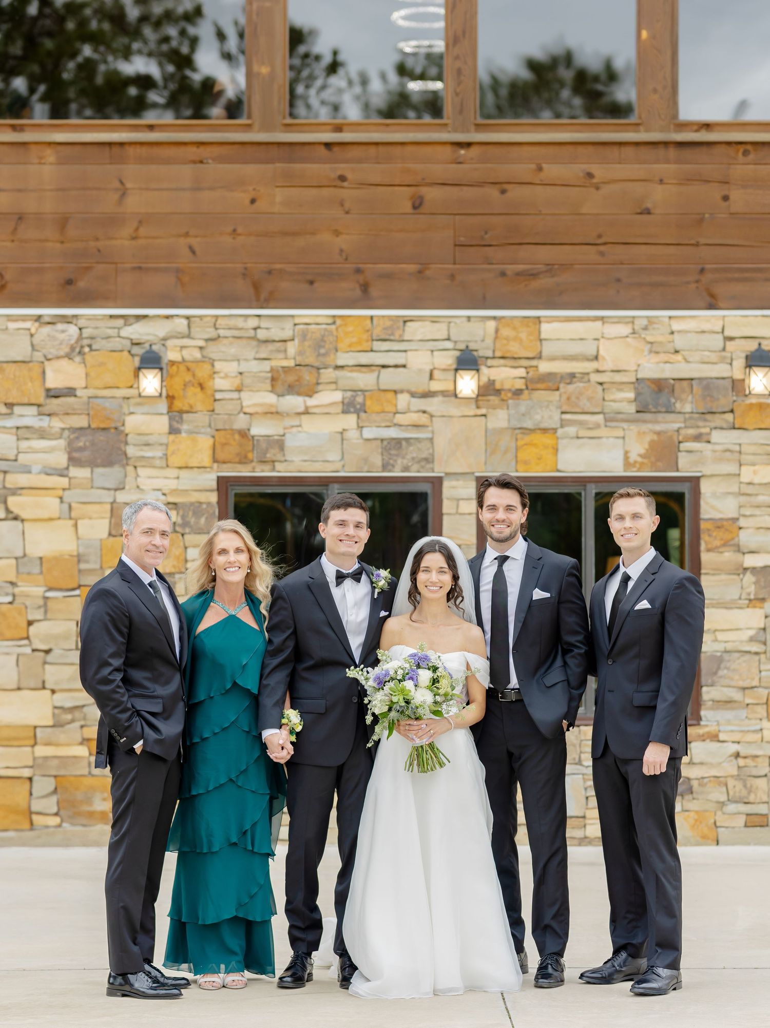 Wedding party poses together in front of stone wall wearing formal attire with black suits and emerald green dresses.