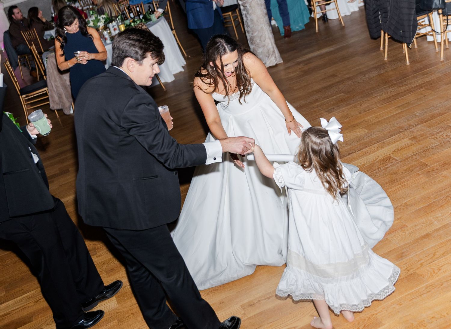 Bride and groom dancing with flower girl in white dress at elegant wedding reception on hardwood floor.