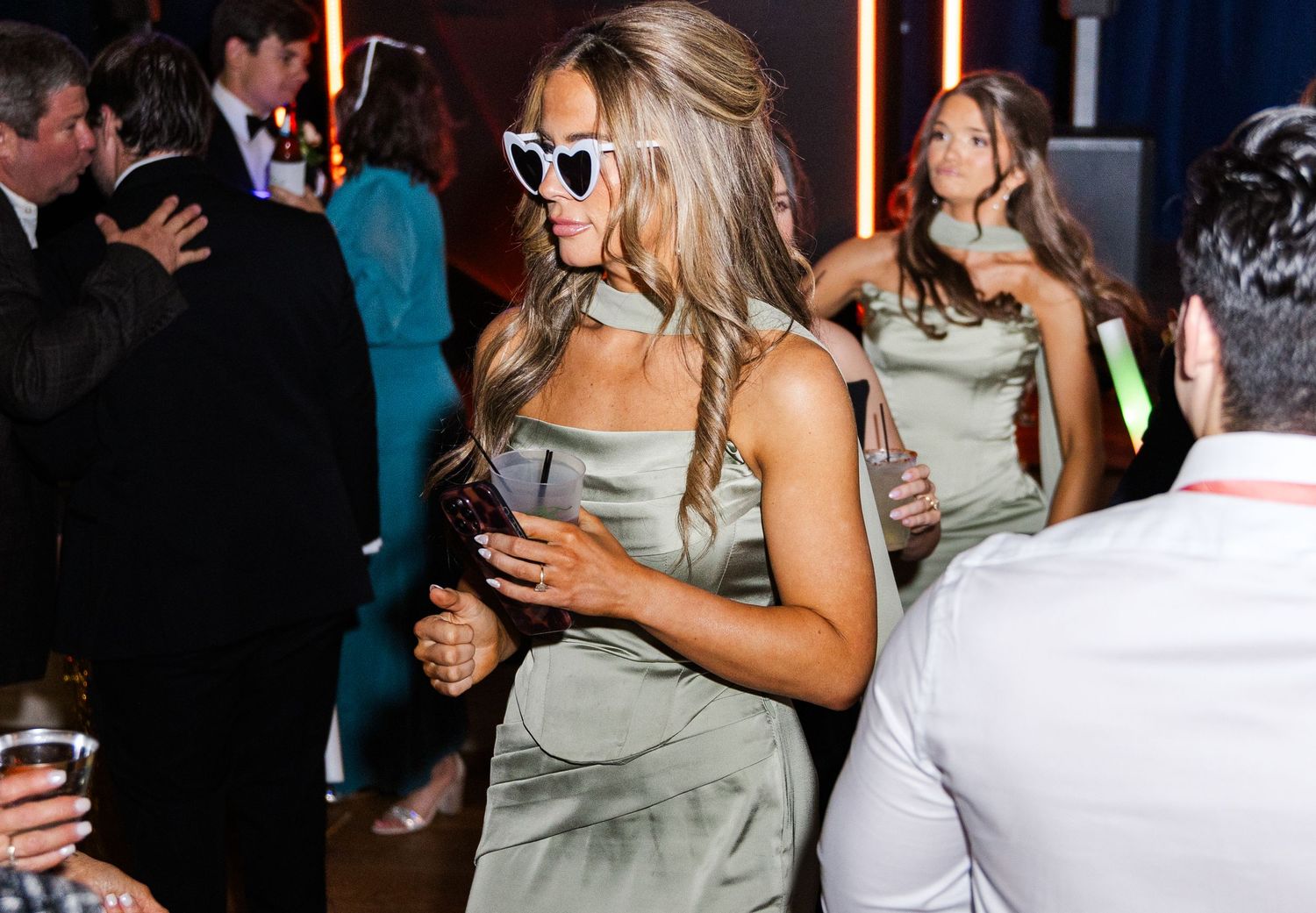 Woman in sage green dress dancing with heart-shaped sunglasses at lively wedding reception.