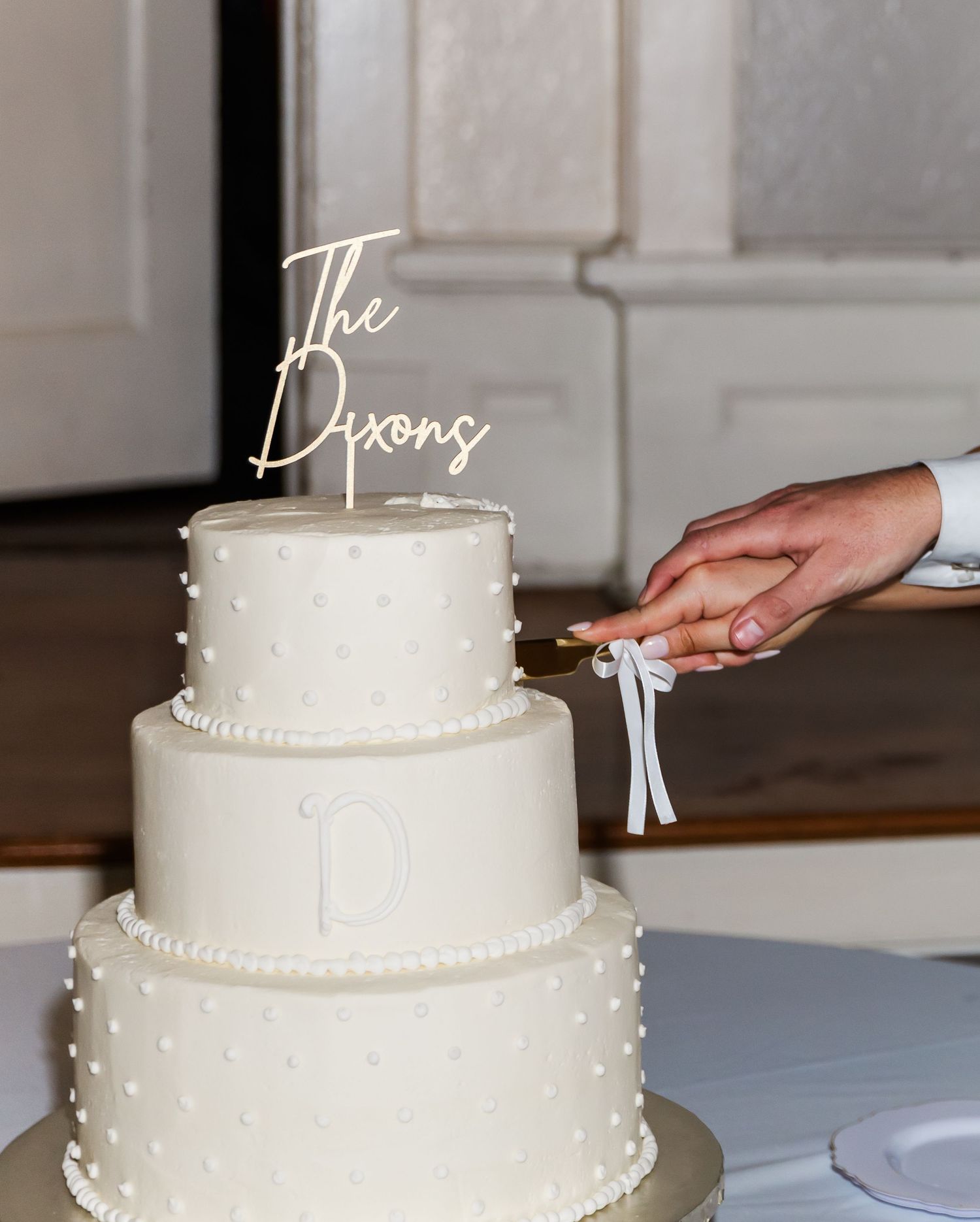 Two-tier white wedding cake with “The Dixons” topper and dotted texture, being cut by the couple.
