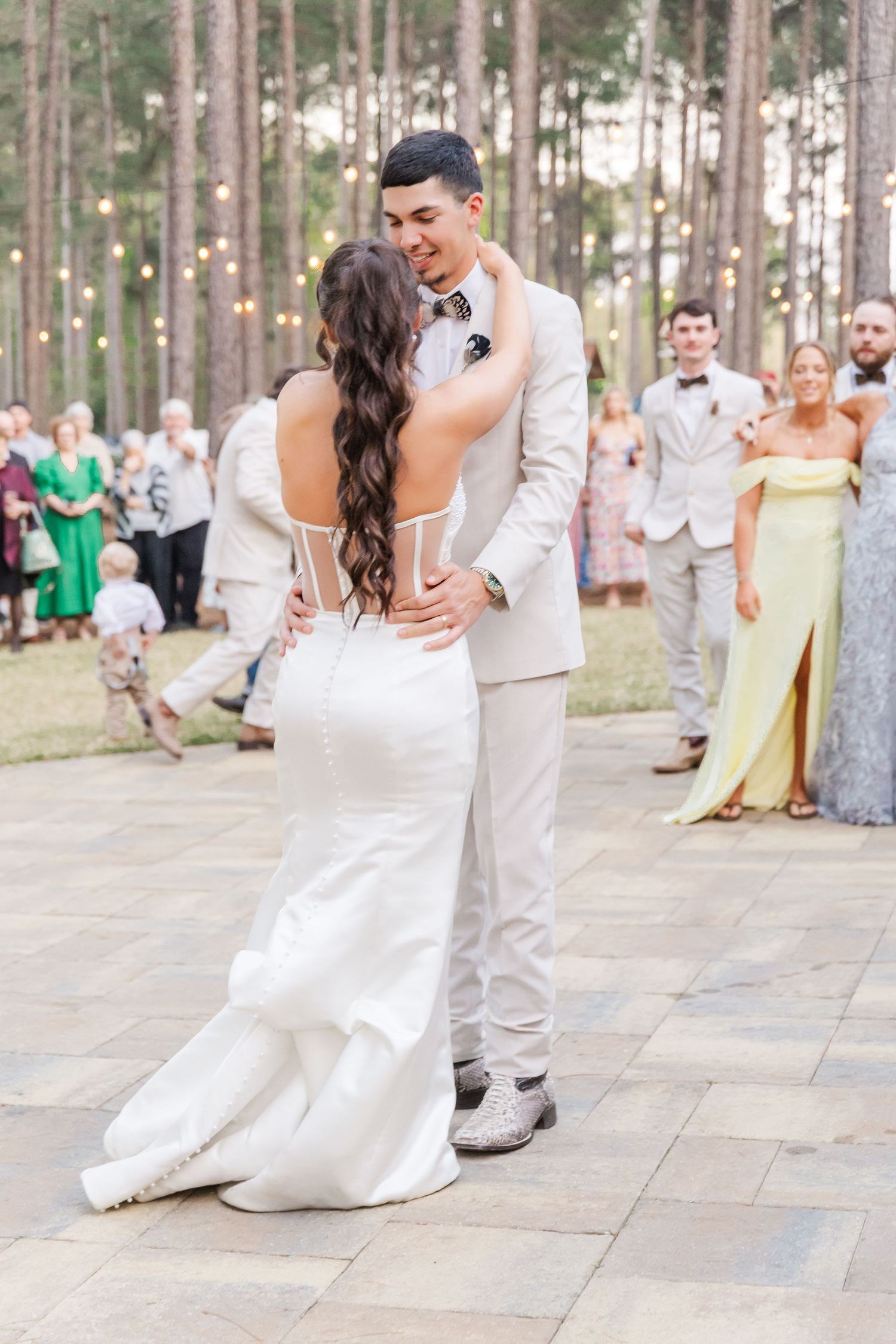 Bride and groom sharing first dance on outdoor patio surrounded by string lights and wedding guests.
