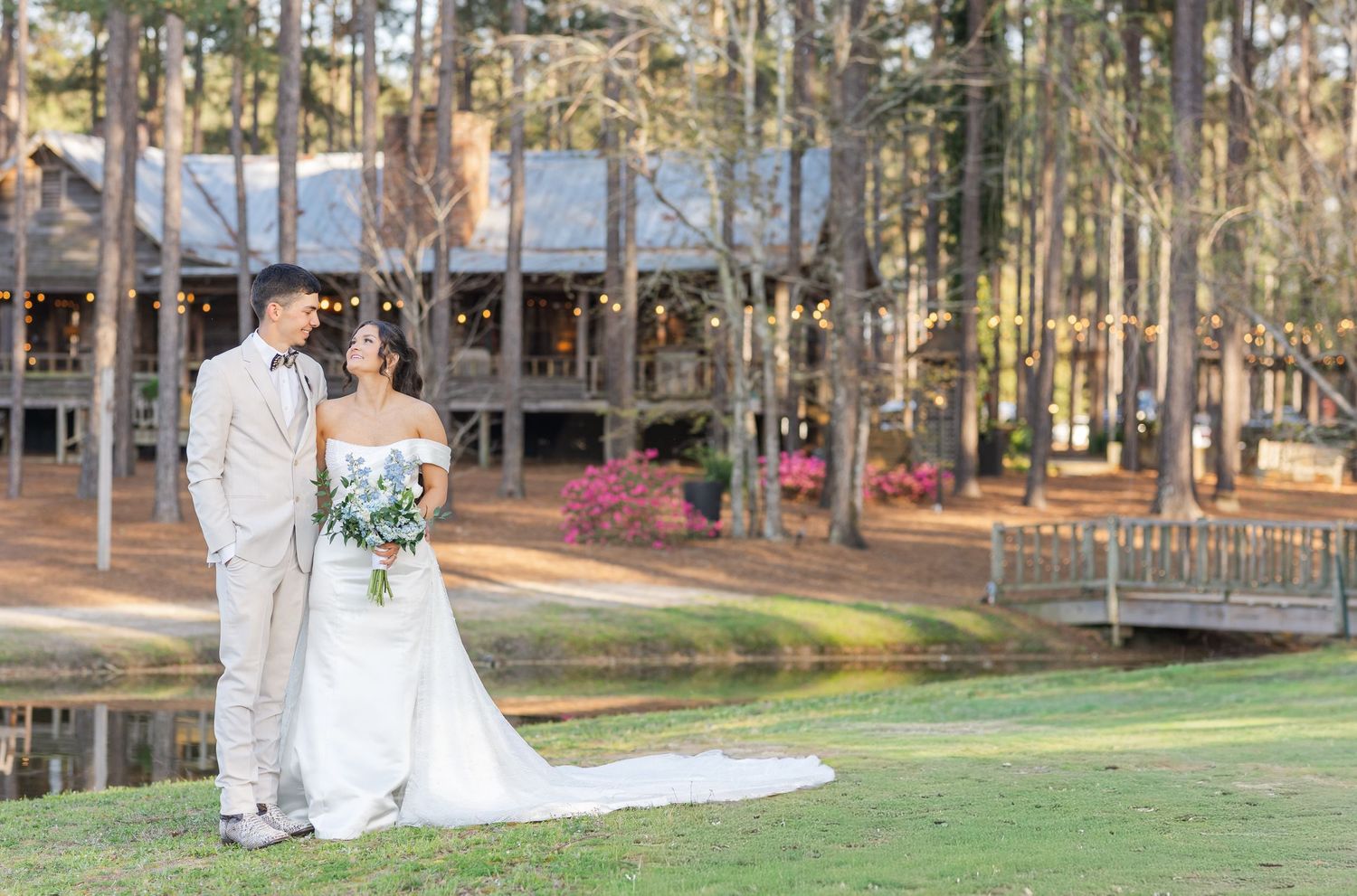 Bride and groom standing on green lawn near pond with rustic wedding venue among tall pine trees.