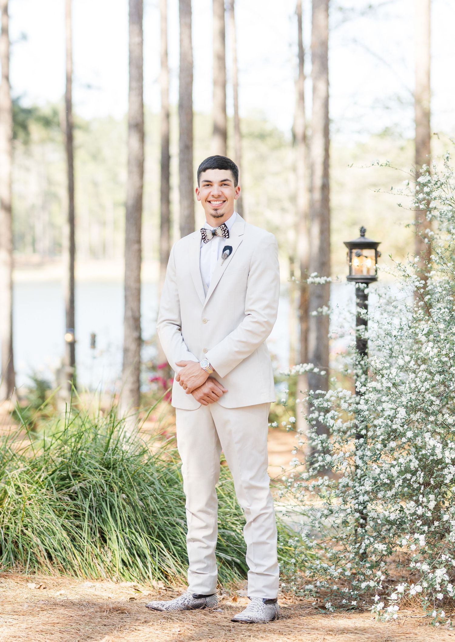 Smiling groom in a tan suit with a floral bow tie standing outdoors among pine trees and white flowers.