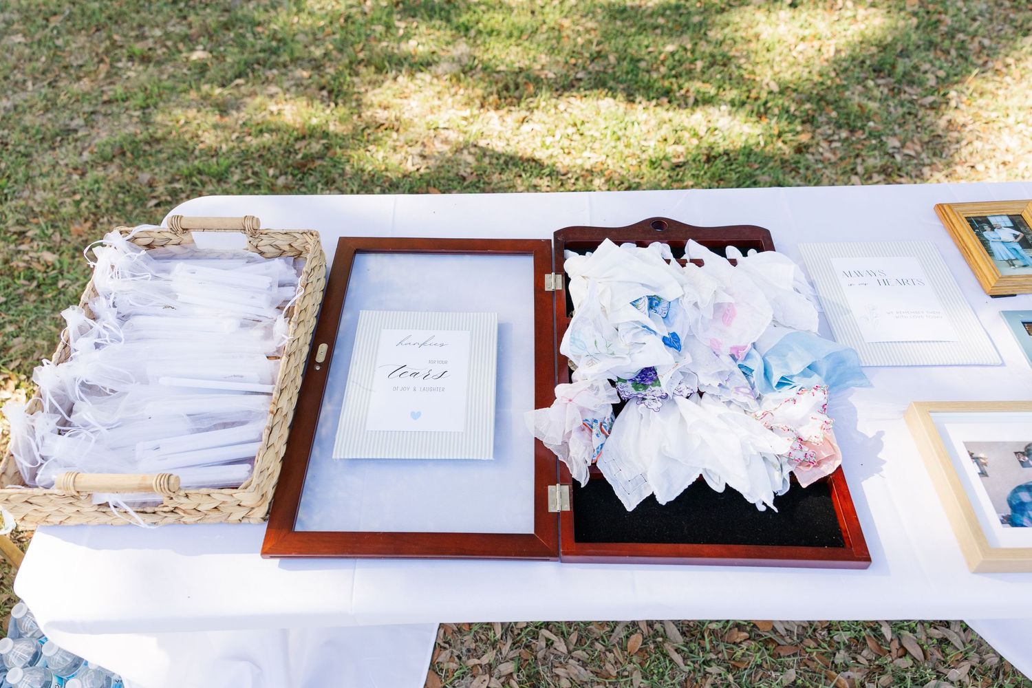 Outdoor wedding welcome table with programs, framed menu cards, and floral fan favors displayed on a white tablecloth.