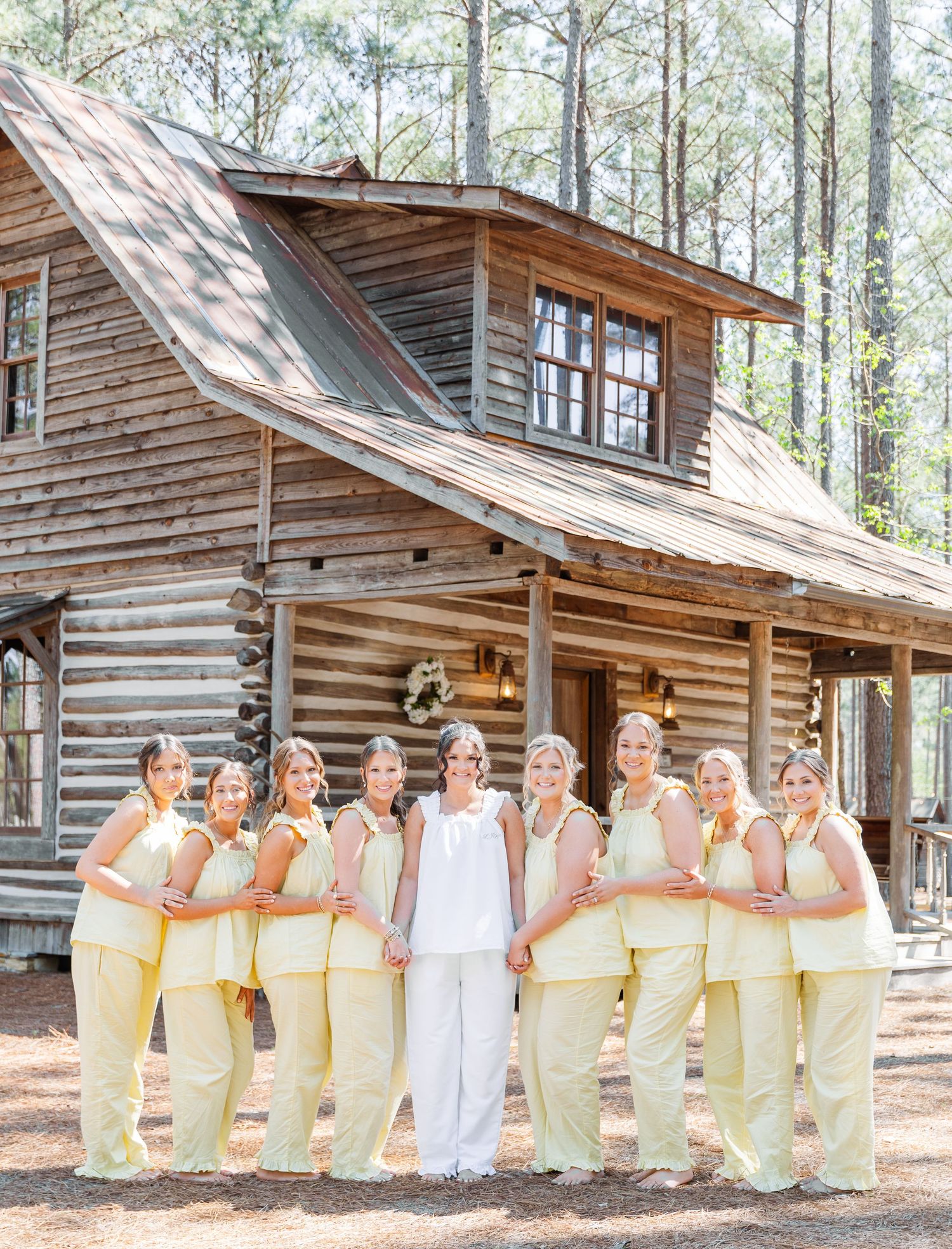 Bride in white stands with bridesmaids in yellow dresses before a rustic log cabin wedding venue surrounded by pine trees.