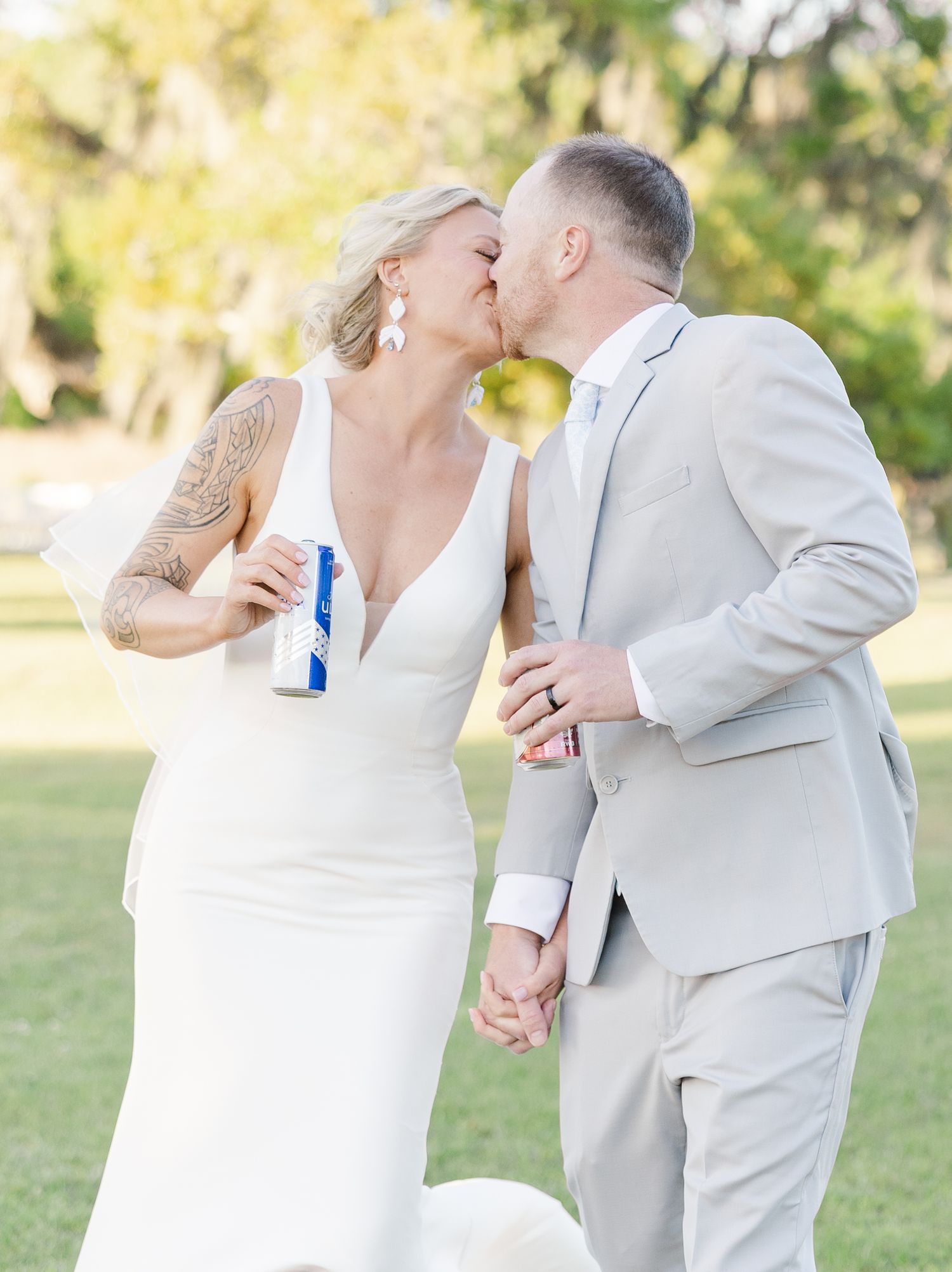Bride and groom share a kiss while holding beer cans on their wedding day outdoors.