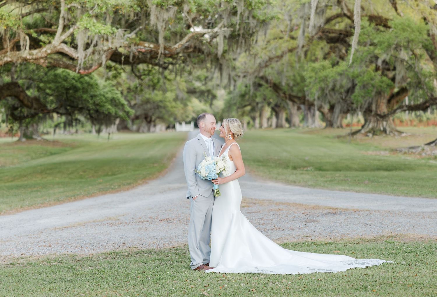 Bride and groom share a kiss on a scenic tree-lined path during their outdoor wedding ceremony.