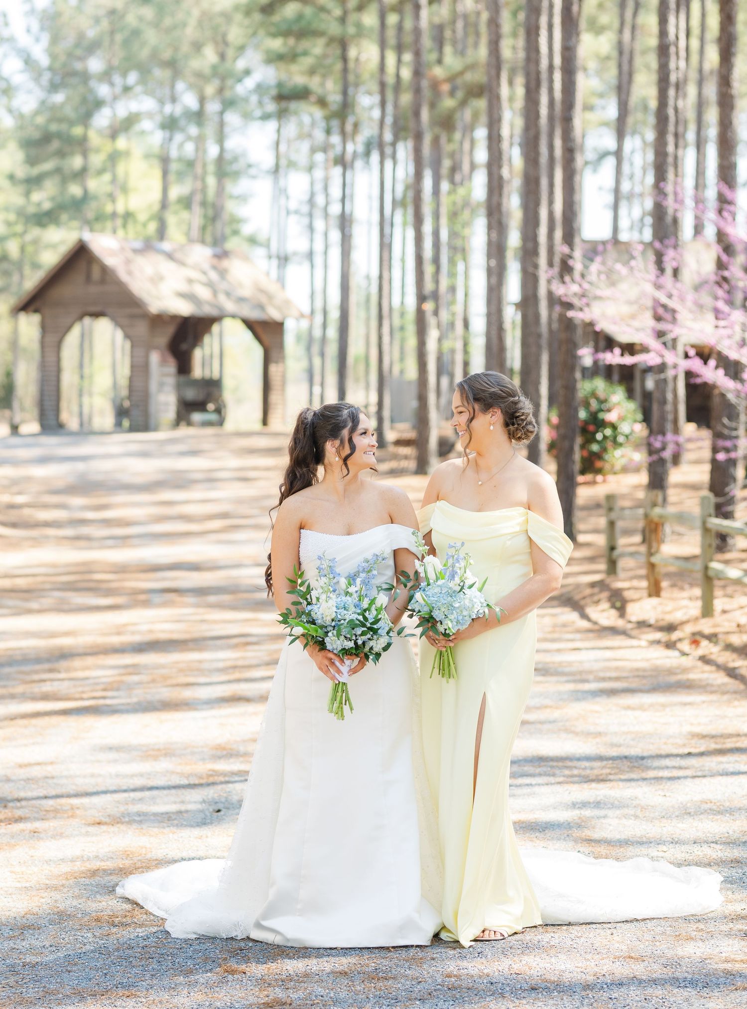 Bride and bridesmaid in yellow gown share a joyful glance while holding blue floral bouquets.