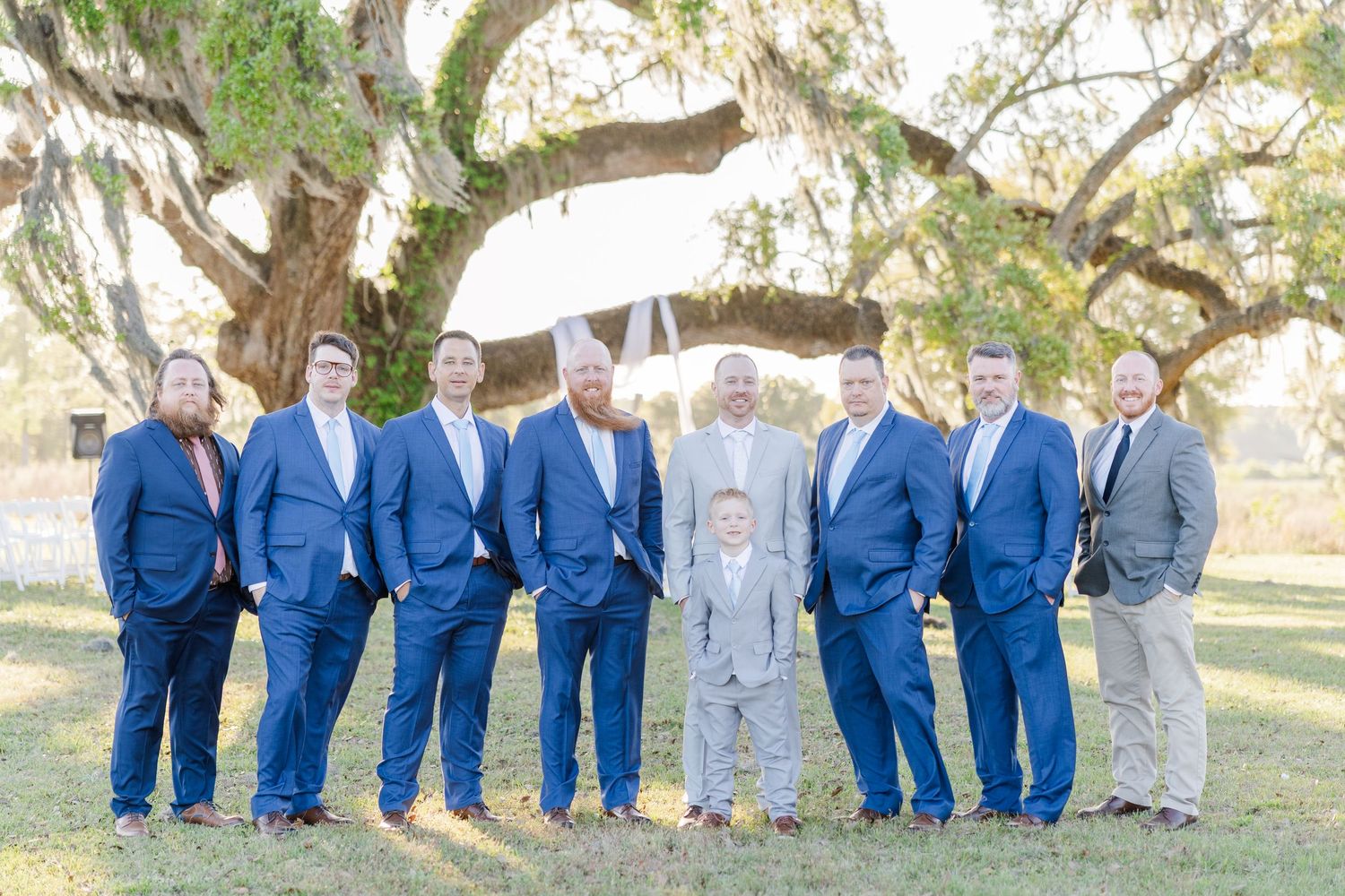 Groomsmen and groom in navy and gray suits posing under a large oak tree draped with Spanish moss.